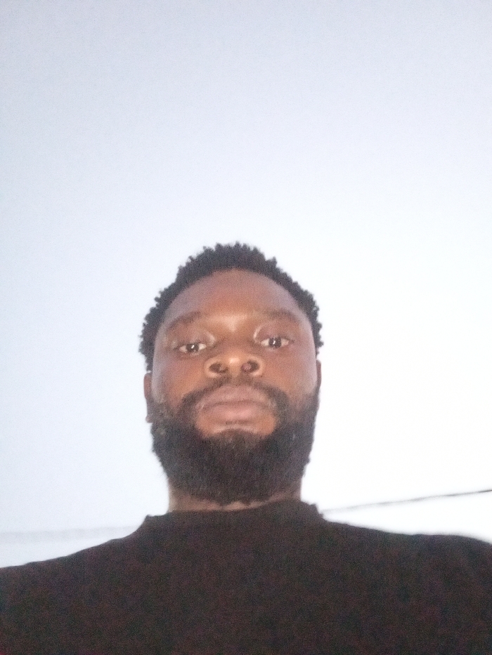 The image displays a close-up, low-angle view of an adult male. He has dark skin, short curly black hair, and a full dark beard and mustache. His facial expression is neutral, and he is looking directly towards the camera. He is wearing a dark, possibly black, crew-neck shirt, with only the upper chest and shoulders visible. The background is a plain, light-colored, undifferentiated surface, likely a wall or an overcast sky, providing no discernible environmental features or geographical markers to indicate a specific location within Magboro, Nigeria, or any other city. No other individuals or objects are visible.