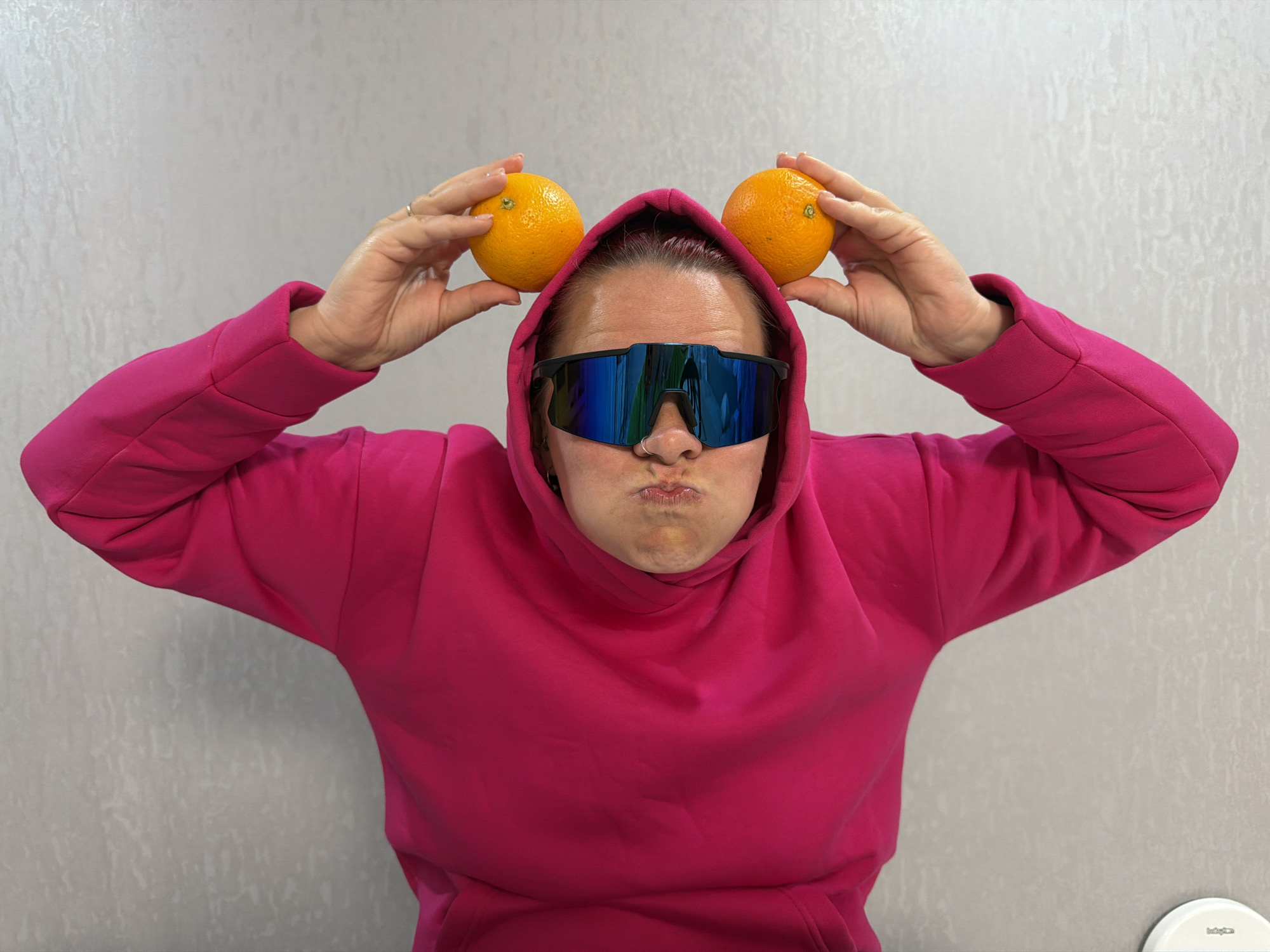 A person wearing a hot pink hoodie and mirrored sunglasses is holding two oranges up to their head, positioned like ears. They are making a puckered expression with their lips. The background is a plain, light grey wall with a subtle texture. A white object is partially visible in the bottom right corner. The location is Unknown, Unknown.