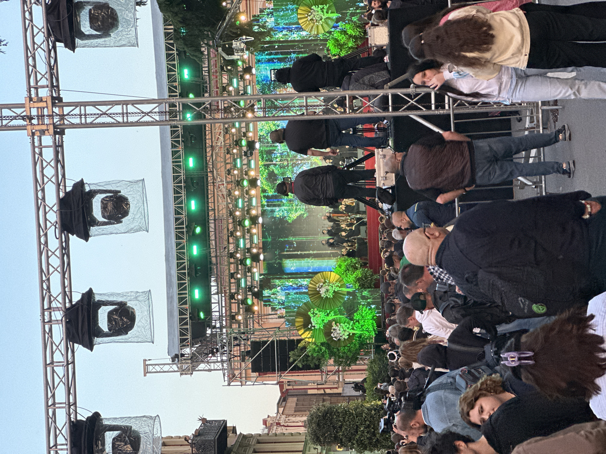 The image shows a public outdoor event, likely a concert or performance, in an urban setting. A large crowd of people is gathered in what appears to be a street or plaza. In the background, there is a stage with elaborate decorations, including what look like oversized green and white flower arrangements. The stage is lit with green lights and what appears to be a video screen displaying forest scenes. A metal scaffolding structure is visible on the sides and above the stage, and there are spherical objects covered in plastic bags attached to the structure. Some people are standing behind a guardrail, and people are sitting in the foreground. The location is unknown.
