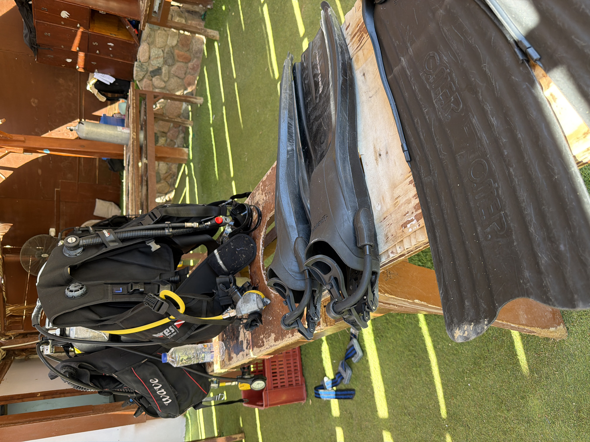 The image depicts scuba diving equipment laid out on a wooden table in an outdoor setting. A black buoyancy compensator (BC) with yellow hoses and regulators is prominent, along with a silver dive cylinder behind it. Two pairs of black swim fins are visible on the table, one pair resting on top of the other. To the left of the BC, a red plastic crate contains a blue and black strap. The table is situated on a green surface that appears to be artificial turf. In the background, wooden lockers and a stone wall are present, suggesting a dive center or rental facility. The overall lighting indicates it is daytime, with sunlight casting distinct shadows. The location is Sharm El Sheikh, Egypt, which is a popular tourist destination known for its diving and snorkeling sites along the Red Sea.