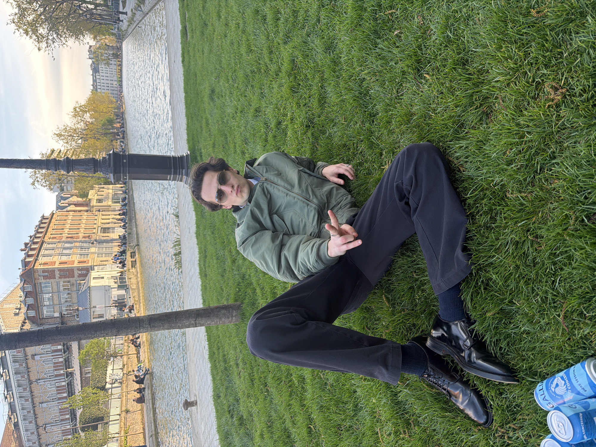 The scene depicts a young man reclining on a grassy lawn in Paris, France. He is wearing a green bomber jacket, dark trousers, black socks, and polished black shoes. He is also wearing sunglasses and has brown hair. He is making a peace sign with his right hand. Several blue and silver beer cans are visible near his feet. Behind him, there is a canal lined with buildings, and a lamp post stands nearby. People are sitting and walking along the canal bank in the background. Trees are visible in the distance. The sky is partly cloudy.