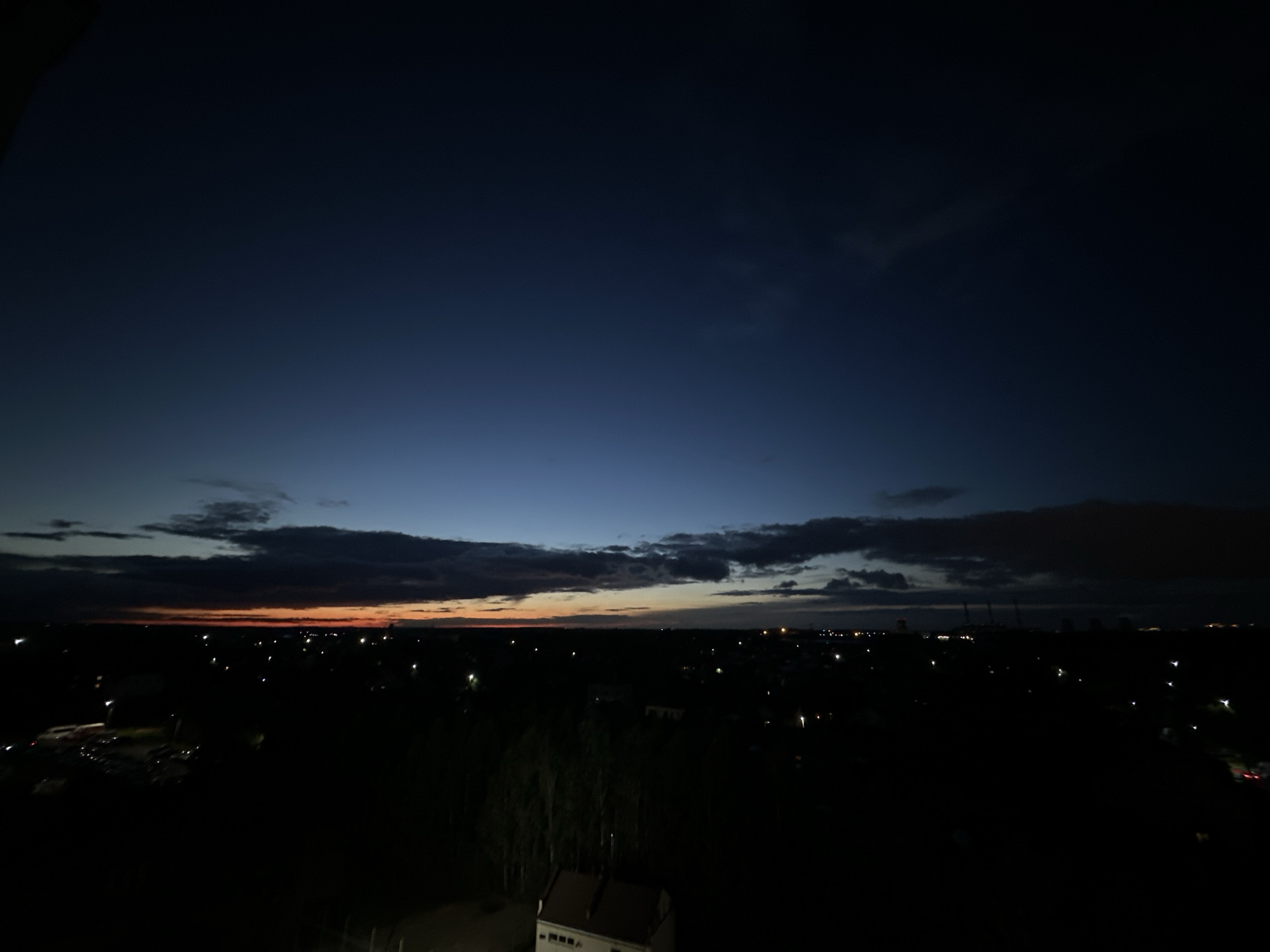 The image depicts a panoramic view of a city at dusk, with the sky transitioning from a deep blue to a faint orange glow near the horizon. Scattered lights from buildings and streetlamps dot the dark landscape, indicating the presence of a populated area. The scene is characterized by the absence of clear details in the foreground and middle ground due to the low light conditions, making specific identification of structures and objects difficult. The overall impression is one of a quiet evening in an urban or suburban environment. The location, Шчомысліцкі сельскі Савет, Belarus, suggests a rural administrative unit that may encompass a settlement or town.