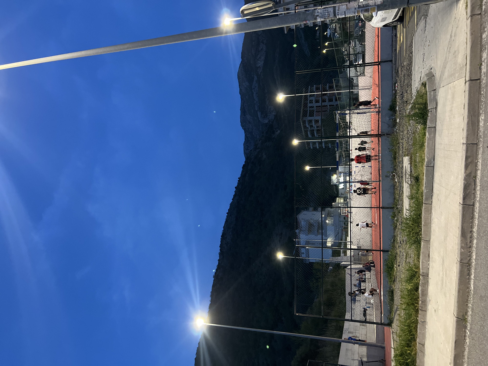 The image shows an outdoor basketball court in Boreti, Montenegro at night, illuminated by streetlights. The court has a red running track around it. A mountain rises steeply behind the court. People are playing basketball and some are sitting on a bench behind the court. There is a concrete barrier next to the court, and a white car is parked near the edge. A tall streetlight dominates the left side of the image. The sky is a dark blue.