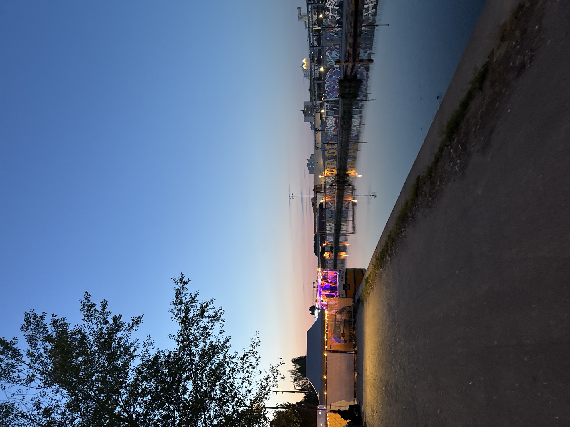 The image shows a cityscape at dusk, seemingly next to a body of water, possibly a canal or river. The photo is taken from a ground-level perspective. The sky transitions from a bright blue to a dusky orange/pink hue near the horizon. Buildings and structures are visible on the opposite side of the water, with lights reflecting off its surface. Some buildings are industrial in nature.
In the foreground, a path or walkway runs alongside the water, and a tree with dark foliage is partially visible on the left side. A temporary structure, potentially a food or beverage stand with lights, is also visible near the path. The lighting suggests it is either late evening or early morning. There are no clear signs of people in the immediate scene, but the temporary structure suggests possible human activity. The location of the image is Paris, France.