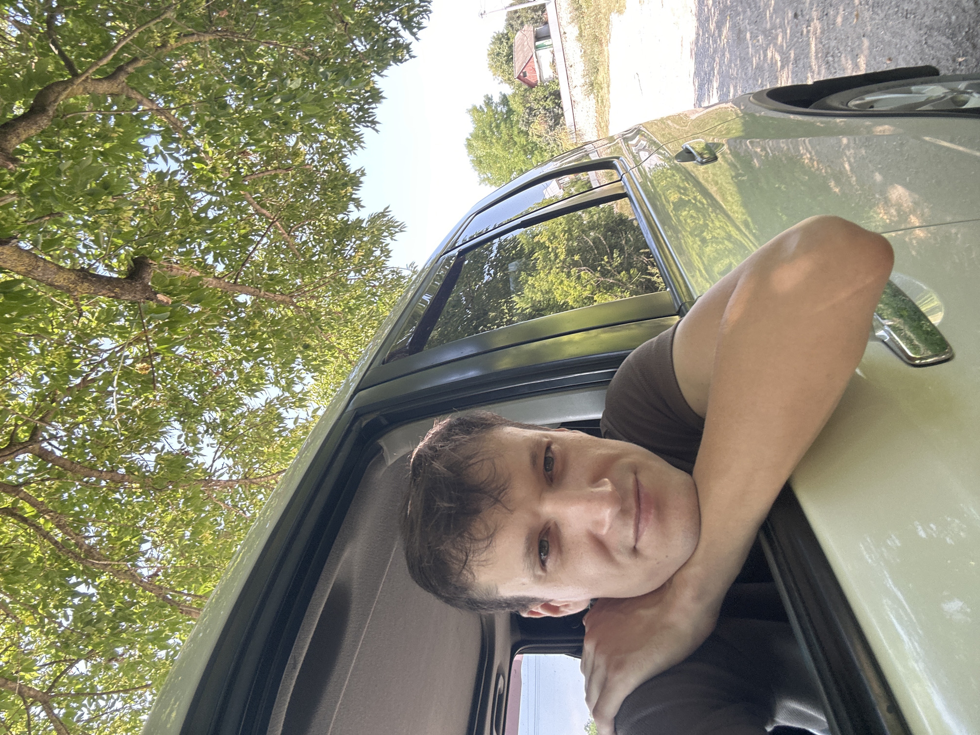 A man is leaning out of a car window, resting his chin on his crossed arms. He is wearing a brown t-shirt. The car appears to be parked on a dirt road next to a grassy area. Trees with green leaves are overhead, casting shadows. In the background, there are houses and more trees. The sky is clear and blue. The overall impression is a casual outdoor scene.