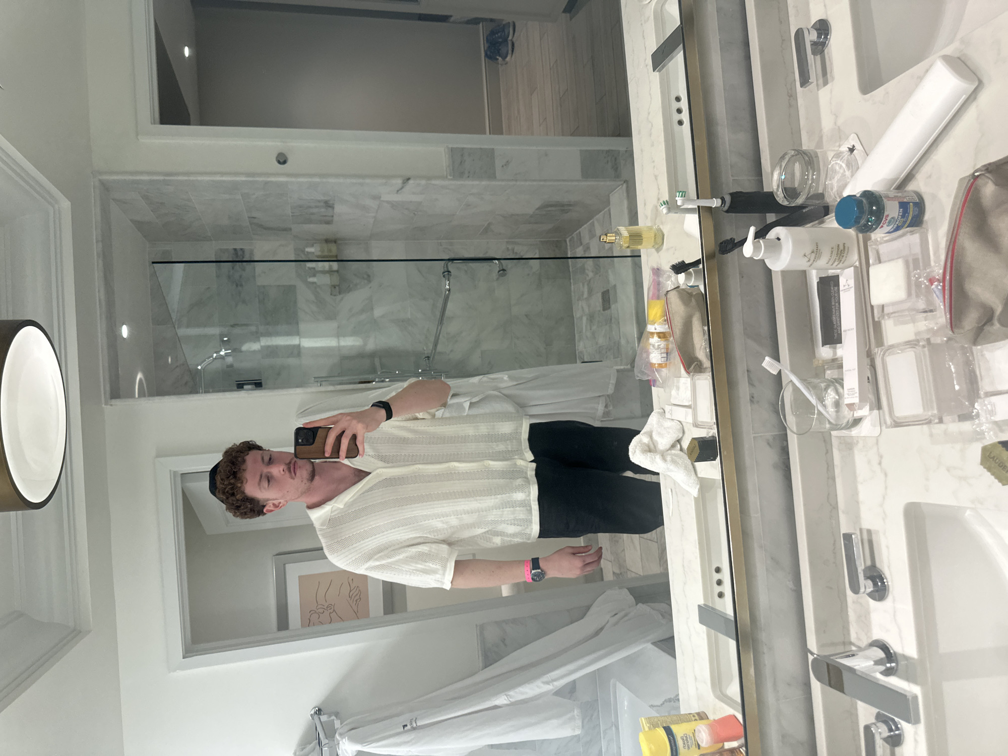 The image depicts a man taking a mirror selfie in a bathroom, likely in a hotel room. The bathroom has a modern design with white marble countertops, a double sink, and chrome fixtures. There is a glass-enclosed shower in the background with marble tile walls. Various toiletries are arranged on the counter. The man is wearing a white collared shirt and black pants. He is holding a smartphone up to take the photo. Based on visual cues, the location is likely in Aventura, United States of America, as Aventura is known for its upscale hotels and modern architecture.