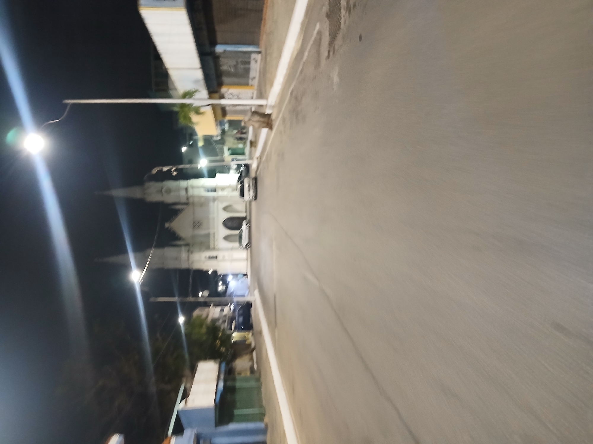 The image shows a street in Juan Griego, Venezuela, at night. The road stretches into the distance with white lines marking the edges. A church is visible in the background. Streetlights illuminate the area, creating bright spots along the road. Parked cars are visible along the side of the road near the church. Buildings line the street.