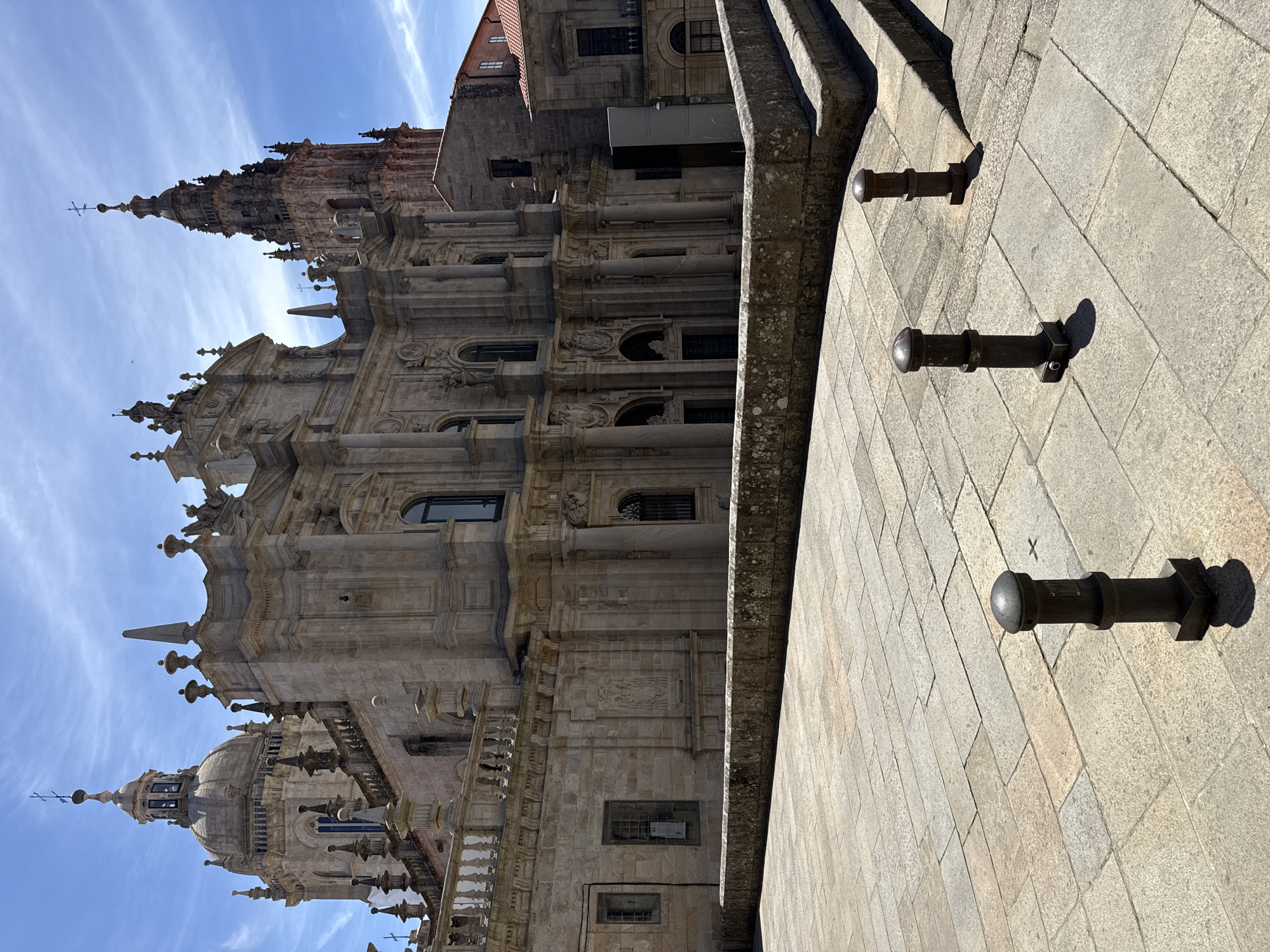 The image captures a section of the Santiago de Compostela Cathedral in Spain, seen from an oblique angle. The architectural style is elaborate Baroque, characterized by intricate stonework, columns, and ornate carvings. A towering spire dominates the left side of the frame, reaching towards a bright blue sky dotted with wispy white clouds. Another dome-like structure with a smaller spire is visible lower down on the left.
In the foreground, a stone-tiled pathway runs diagonally, featuring three dark, cylindrical bollards spaced along its length. The bollards are functional objects, likely serving as traffic or crowd control, and cast distinct shadows on the sunlit pavement. The stone facade of the cathedral exhibits a rough texture and is weathered, suggesting its age. The overall scene is bathed in clear, bright sunlight, indicating it is likely daytime, possibly late morning or early afternoon given the angle of the shadows.
The image evokes a sense of grandeur and historical significance. There is no visible text in the image. The focus is on the impressive architecture and the interplay of light and shadow on the stone surfaces. No people are visible in the frame. The weather appears clear and pleasant.