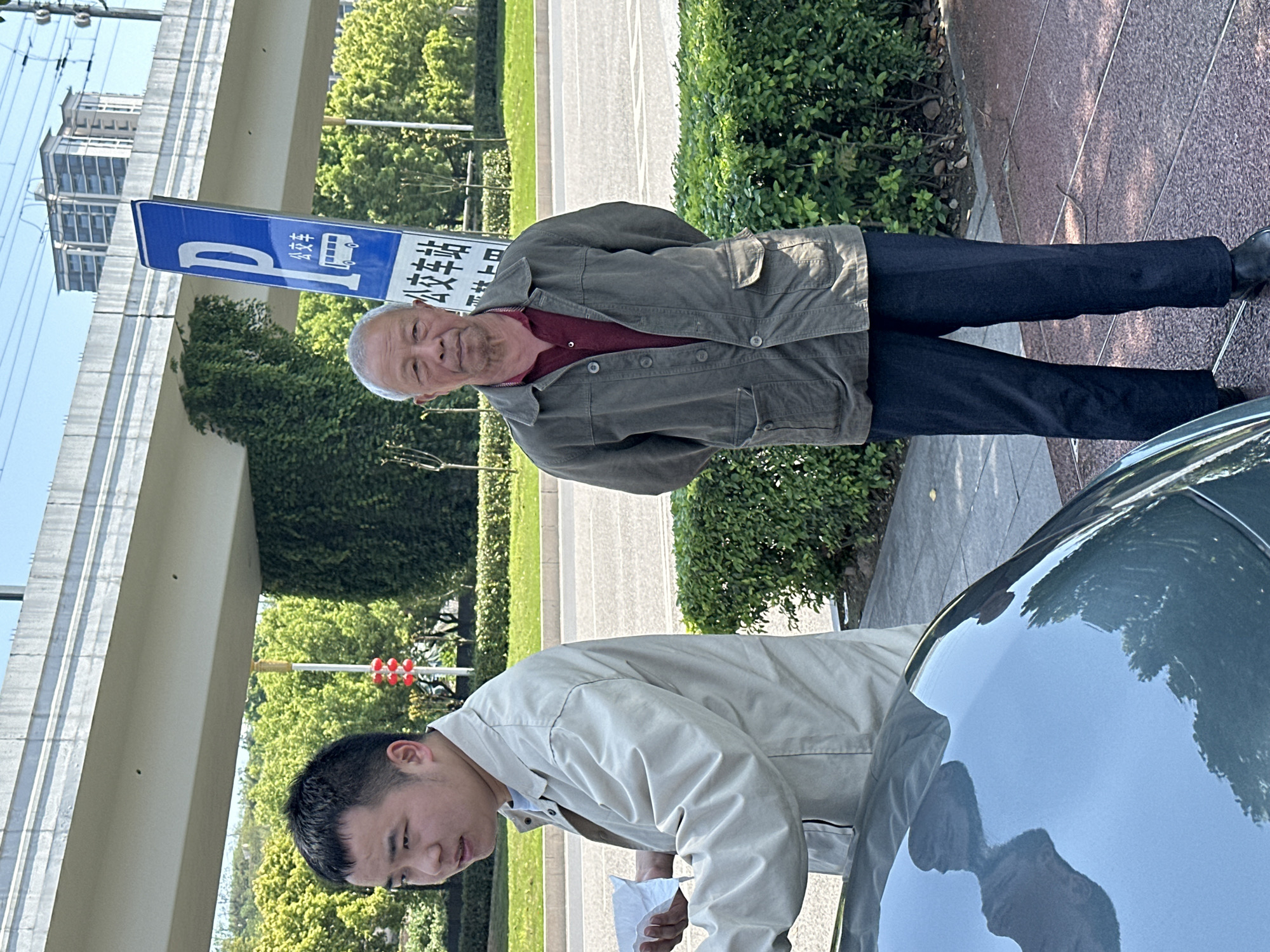 An outdoor daytime shot depicts two men standing near a car on a street in what appears to be an urban environment, possibly Jiangjin, China. The older man, with gray hair and wearing a green jacket over a red shirt and dark pants, is looking at the camera with his hands behind his back. A younger man is seen closer to the car, dressed in a light-colored jacket and holding a piece of paper. A blue parking sign with Chinese characters is visible in the background. The car’s hood is prominently featured, reflecting the surrounding environment. Bushes and a building structure provide additional background detail.