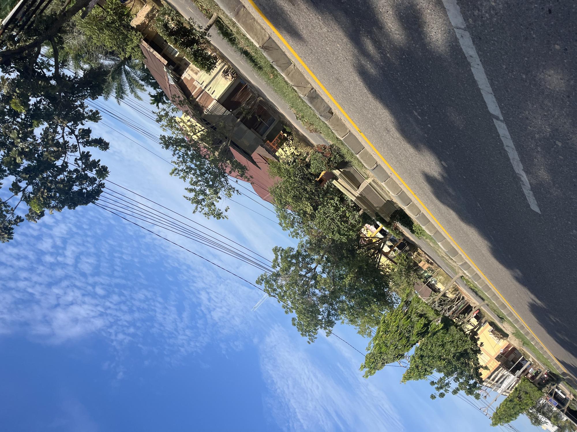 The image depicts a street scene in Tanahterban, Indonesia, on a clear day with a blue sky and scattered clouds. A paved road runs diagonally across the frame, with a yellow line marking the edge of the lane. A curb with a stone pattern separates the road from a grassy verge.
Along the right side of the street, there are several buildings with corrugated metal roofs and light-colored walls, some of which appear to be older structures. Trees and lush green foliage line the street in front of the buildings, with some taller palm trees visible further in the background. Power lines are strung across the sky, supported by poles that are not fully visible. A faint contrail from an aircraft is visible in the upper left quadrant of the sky. The perspective suggests the photo was taken from a moving vehicle or at an angle looking down at the road.