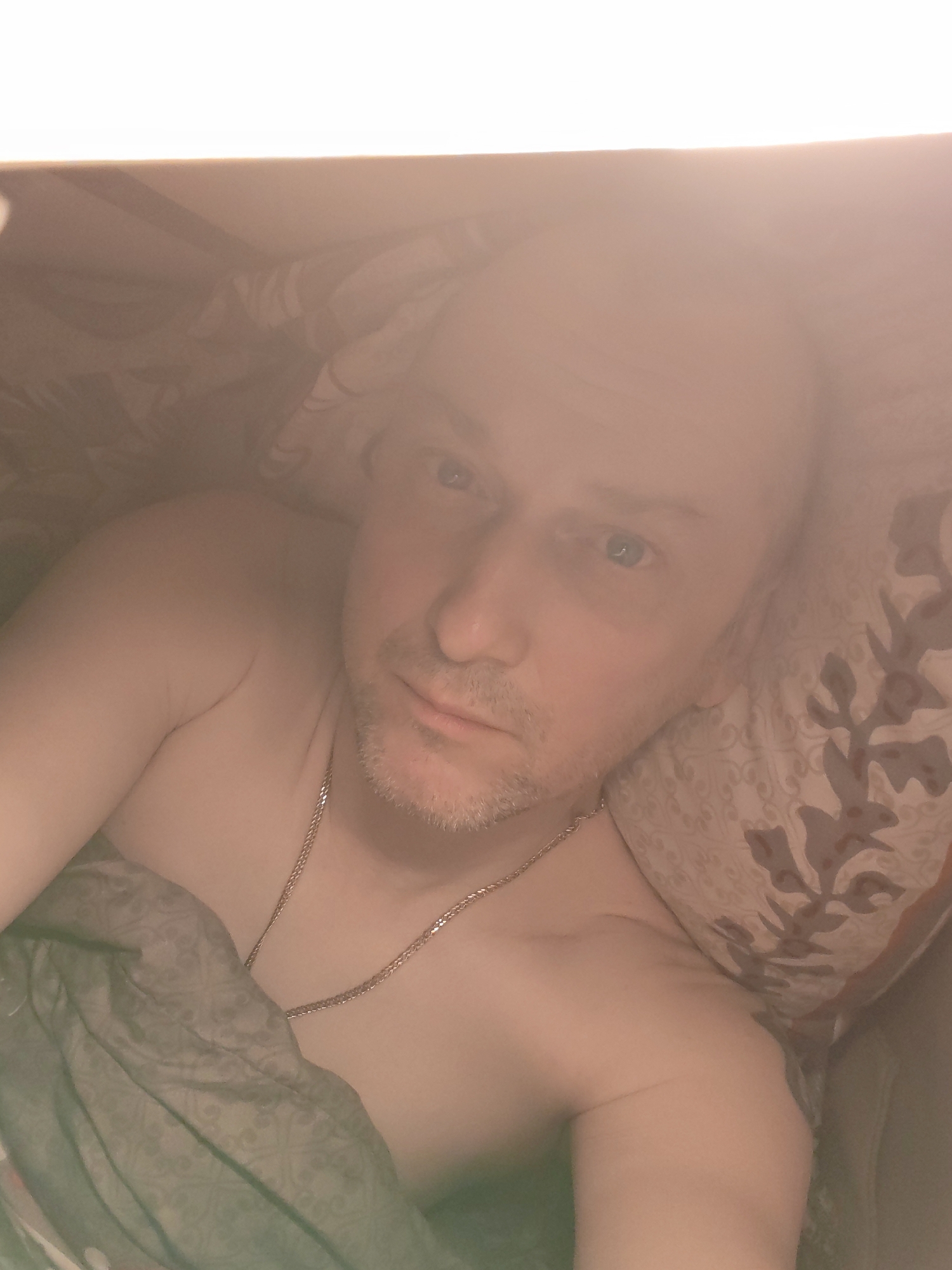 A man with light skin and blue eyes lies in bed. He has a shaved head and light stubble. He is wearing a thin silver chain around his neck. A gray patterned blanket covers part of his chest. His head rests on a patterned pillow. The setting appears to be indoors, possibly in a residential location. There's no information to confirm that the photo was taken in Bol'shie Mkhi, Russia.