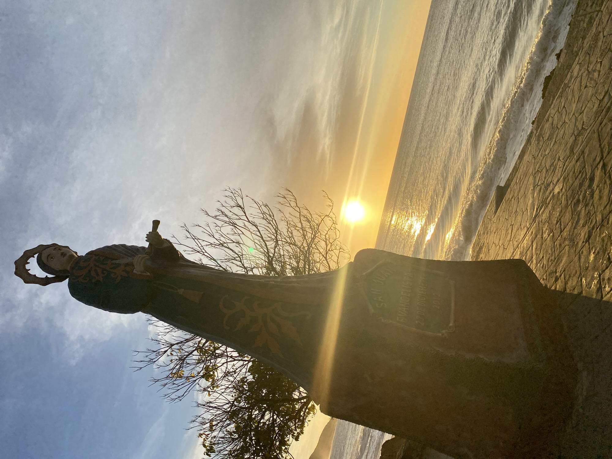 The image shows a statue of a saint with a halo on a pedestal, silhouetted against a sunset at the beach in Juan Griego, Venezuela. The beach is to the right, with waves and the sun reflecting off the water. The statue has foliage in front of it. The pedestal has a plaque with the words "San Juan Evangelista 27 de Diciembre 2017". The sky transitions from pale blue to yellow near the sun. The beach to the right is walled off by cobblestone.