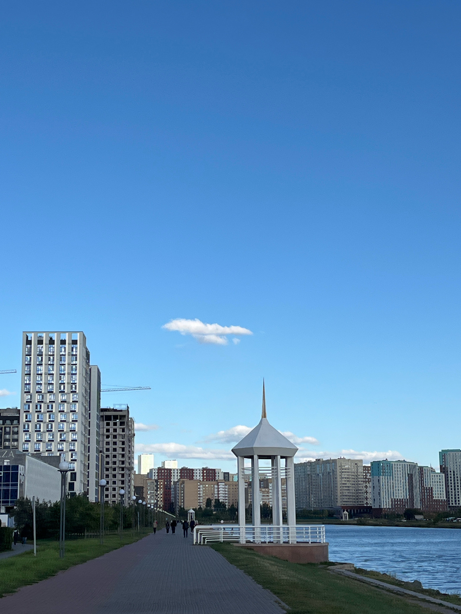 This is a photograph taken in Astana, Kazakhstan, likely during the daytime. The image captures a wide view of a waterfront area with a paved walkway in the foreground. Several modern high-rise buildings are visible in the background, some appearing to be under construction with cranes present. A white gazebo-like structure with a pointed spire stands near the water's edge. The sky is clear blue with a few scattered white clouds. A calm body of water is to the right of the walkway, and lush green grass borders the path. Several figures are walking along the path, appearing as small silhouettes in the distance.