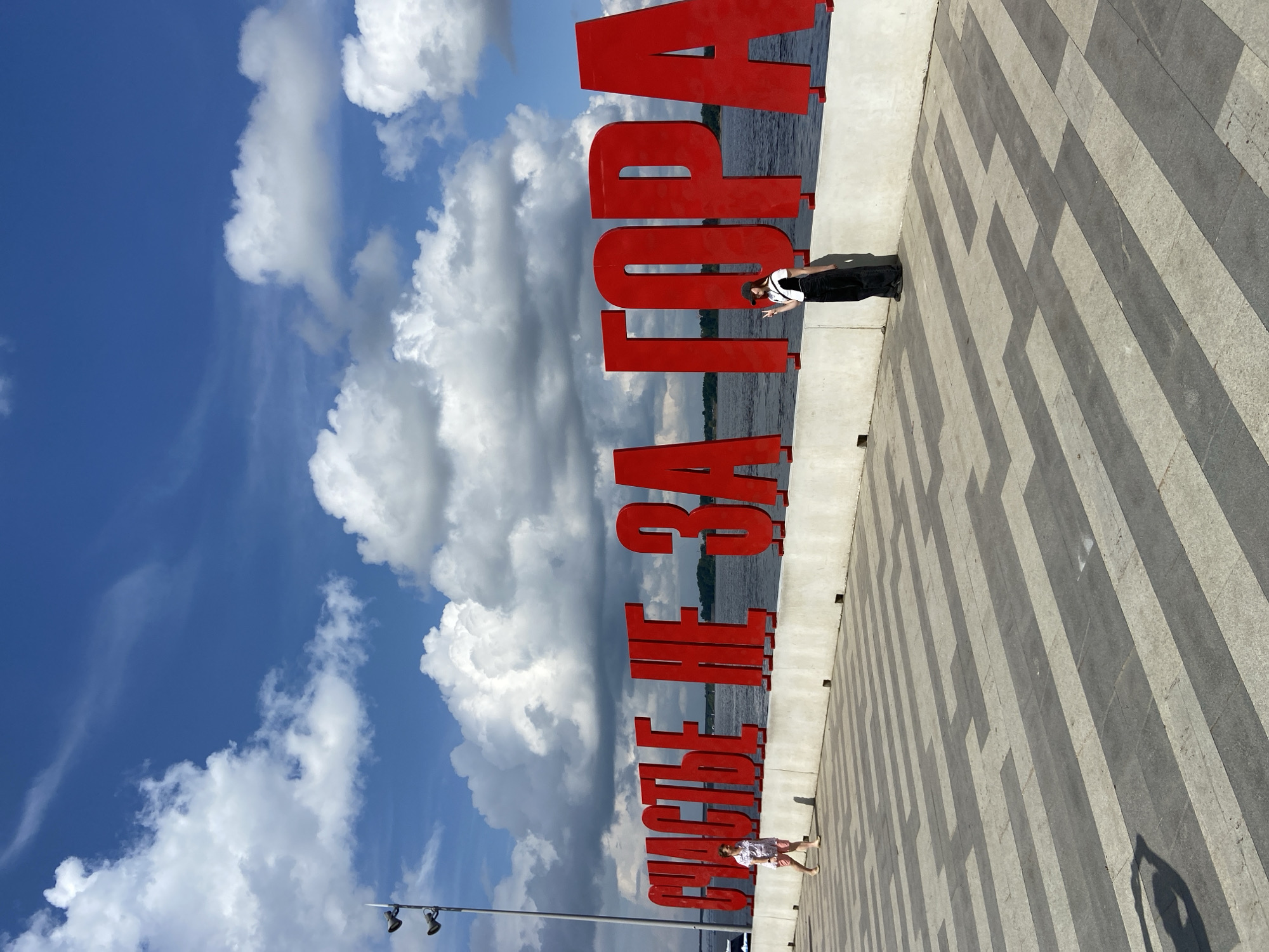 The image shows a waterfront promenade in Perm, Russia, featuring large red three-dimensional letters spelling out "СЧАСТЬЕ НЕ ЗА ГОРОЙ" (Happiness is not far away). The promenade is paved with a pattern of gray and beige rectangular tiles. On the left side of the image, a person in a light-colored shirt and pink shorts walks away from the camera. On the right side, a person wearing a white top and black overalls stands near the letters, making a peace sign with their hand. Behind the letters is a body of water, likely the Kama River, and the sky is blue with white, puffy clouds. A lamppost with two lights is visible on the far left, extending upwards.
