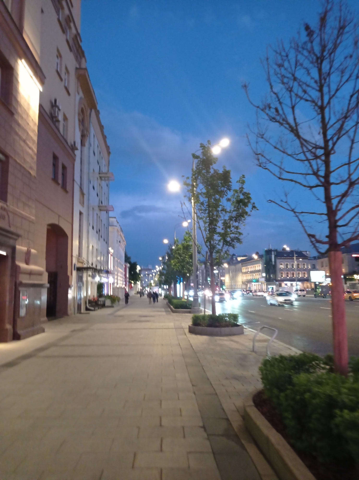 The image depicts a street scene in Moscow, Russia, likely at dusk or early evening. A wide sidewalk paved with light-colored square tiles runs along the left side of the frame, adjacent to the facade of a multi-story building with neoclassical architectural elements. The building features several windows and an arched entrance. Further down the sidewalk, pedestrians are visible walking.
Across the street, a road with multiple lanes of traffic is illuminated by streetlights and the headlights of moving vehicles. Several cars are visible on the road, some appearing to be in motion, with their lights creating streaks of light. Buildings with illuminated windows line the opposite side of the street, suggesting a commercial or residential area. Trees, some with bare branches and others with green foliage, are present along the sidewalk and median. The sky above is a deep blue with some wispy clouds. The overall impression is that of an active urban environment at the transition from day to night.