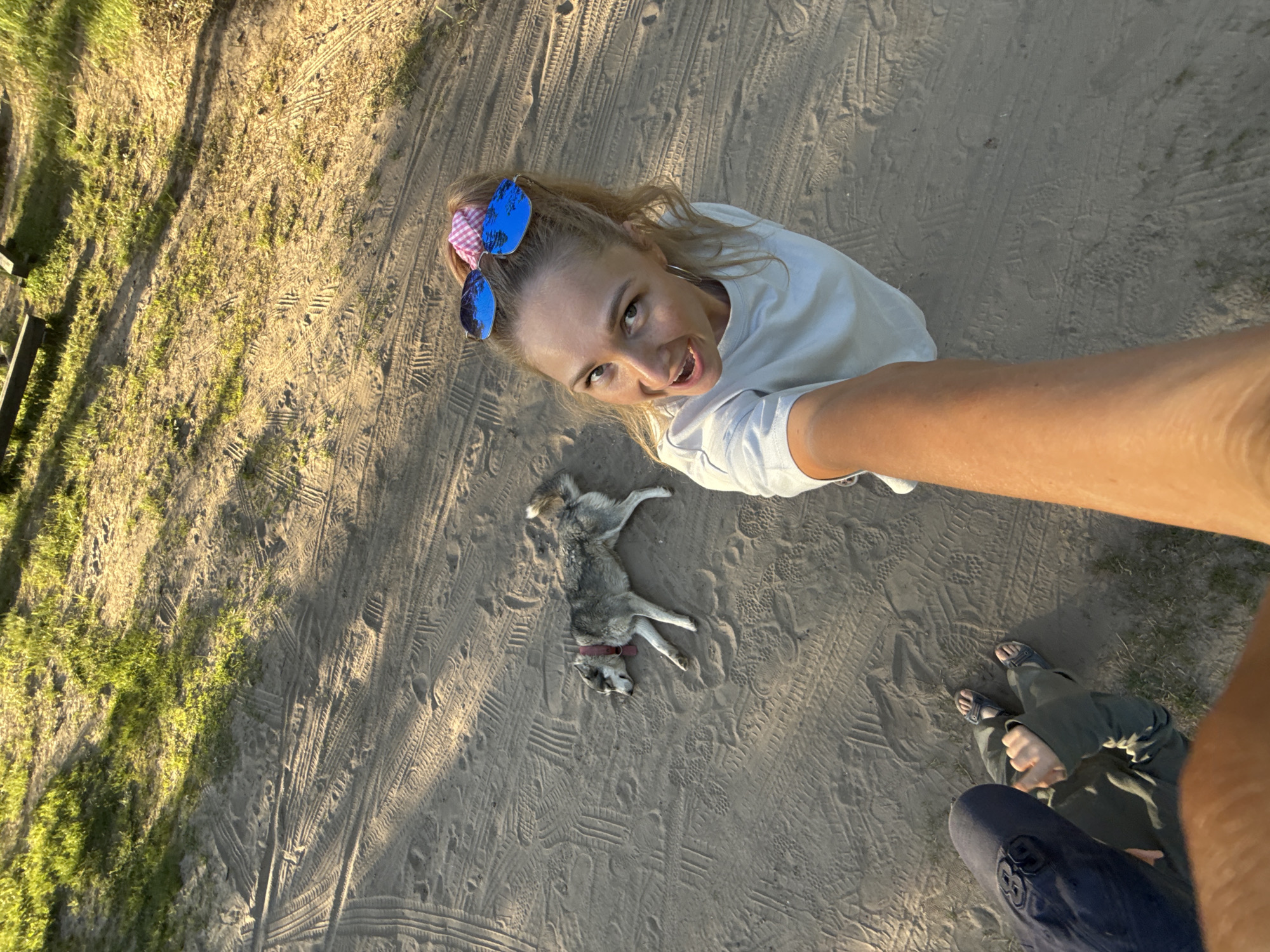 The image shows a woman taking a selfie outdoors. She is wearing a white t-shirt, and blue sunglasses are propped up on her head. She has light brown hair and is smiling at the camera. A grey and white husky dog is lying on the ground next to her. In the background, there is sandy ground with tire tracks and footprints. Green foliage is visible on the left side of the frame. In the bottom right corner, a person's legs and feet, clad in sandals and pants, are visible. Based on the location, the scene may be in Novyi, Russia, but there are no discernible landmarks or signs to confirm it.