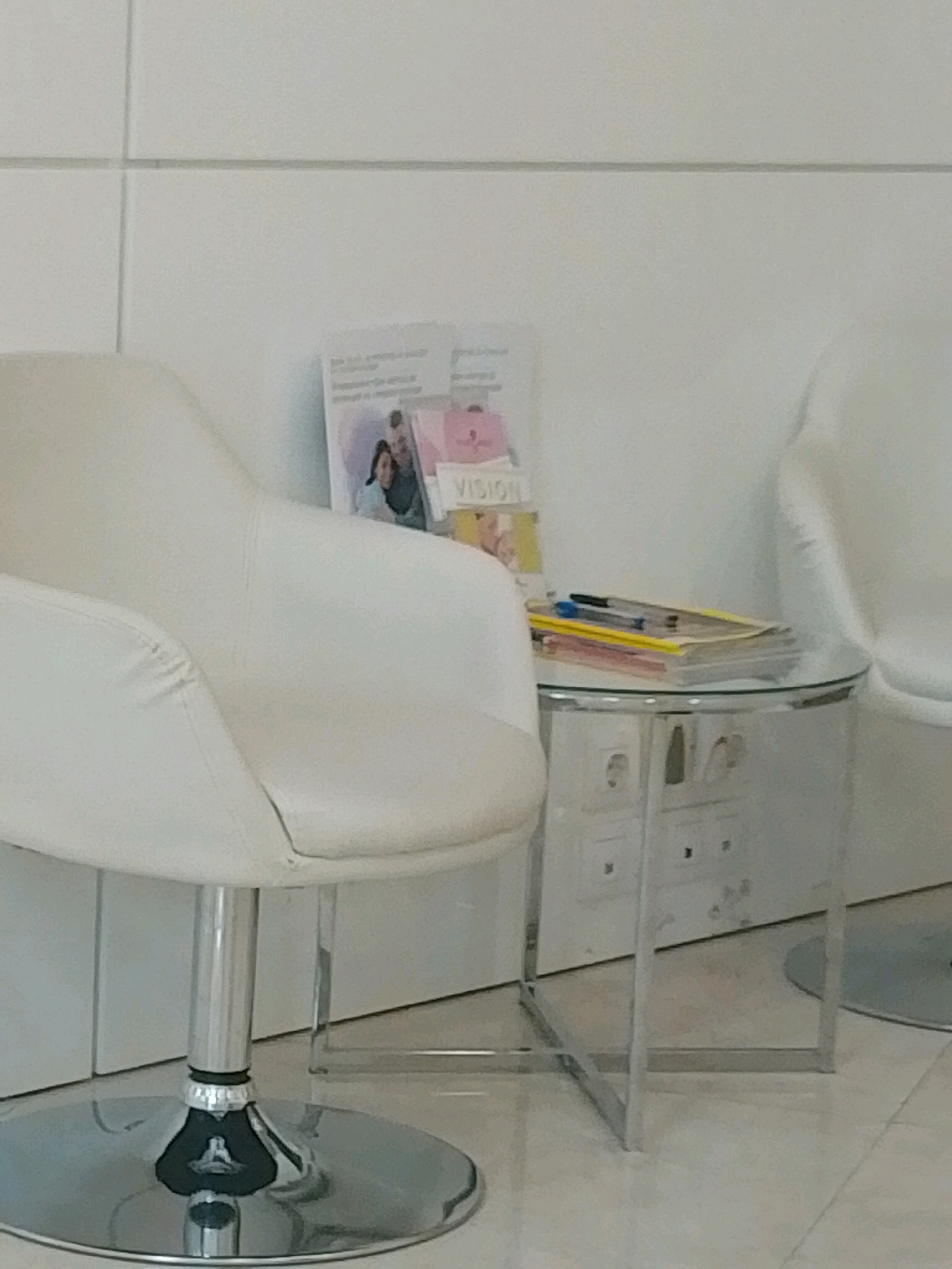 The image shows a waiting room with a white chair and a glass-top side table. On the table, there are a stack of magazines or brochures and two pens. The wall behind the furniture is white and tiled. The floor appears to be light-colored tile. The overall scene suggests a clean and modern waiting area, likely in a professional setting such as a doctor's office or clinic. The exact location within Sofia, Bulgaria, cannot be determined from the image alone.