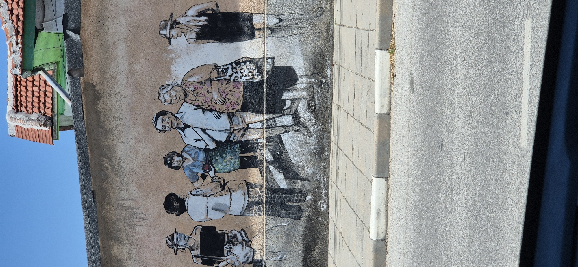 The image shows a mural painted on a building's exterior wall, depicting six figures in what appears to be a street scene. The mural is rendered in black and white with some light color accents on the clothing of some figures. From left to right, the figures include a person wearing a hat, a t-shirt, and shorts; a person facing away from the viewer, wearing a light-colored shirt and pants; a person in a blue shirt and a patterned skirt, holding something red; a man in a light blue shirt and shorts, smiling and with his hand on his hip; a woman in a patterned top and dark skirt, carrying a patterned bag; and a person in a black dress and hat, with long hair. The figures are standing on a textured surface that suggests pavement or ground. The wall itself is a light brown, rough texture. Above the mural, the roofline of the building is visible, with terracotta tiles and some green trim. A clear blue sky is above the building. The bottom of the image shows a paved sidewalk and a street. The scene is outdoors, under daylight.