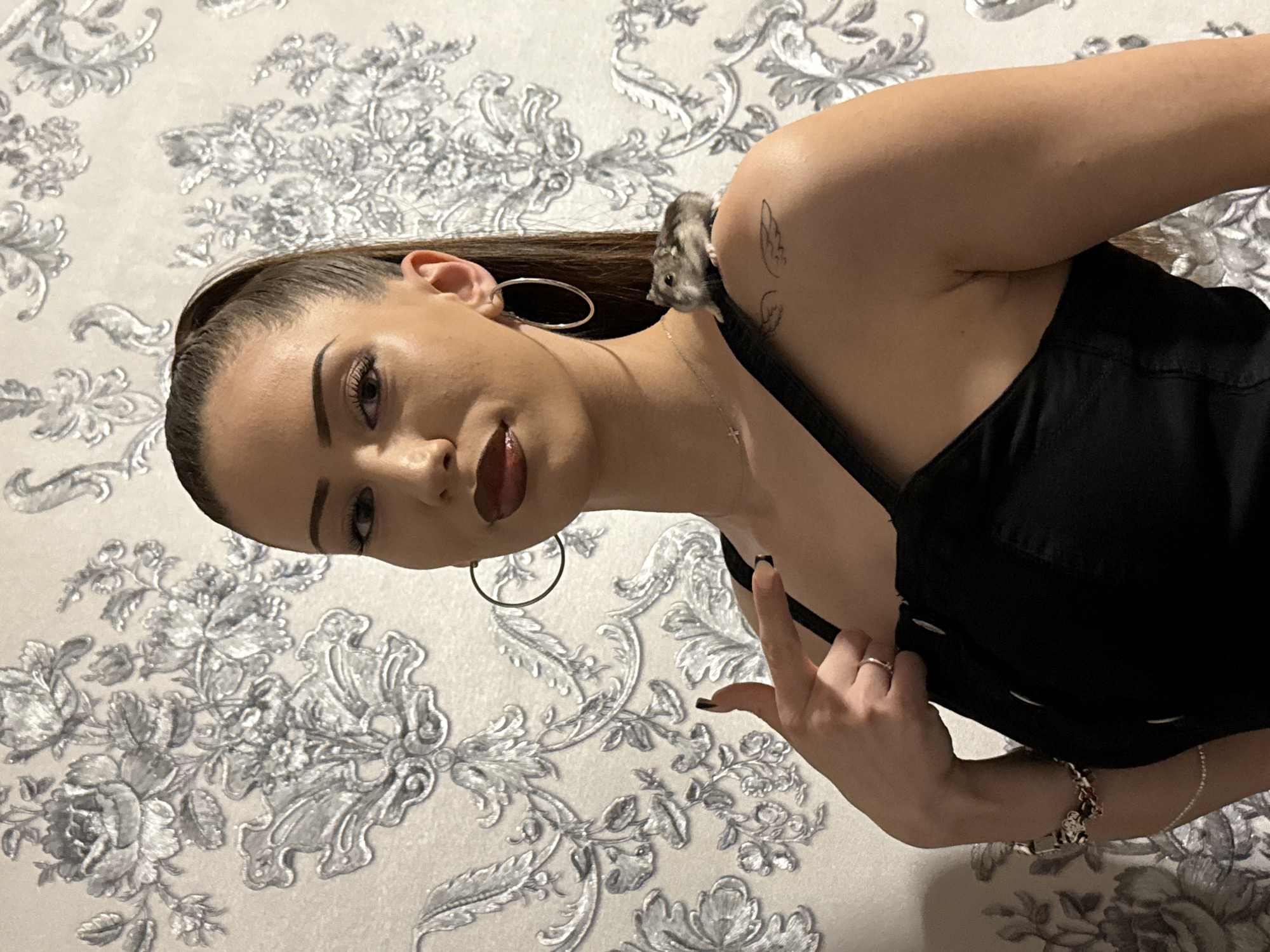A young woman is captured in a medium shot indoors, holding a small rodent on her shoulder. She appears to be in her late teens or early twenties, with dark hair pulled back from her face. She has noticeable eye makeup, including winged eyeliner, and dark red lipstick. She is dressed in a black, sleeveless top or dress, and accessorized with large silver hoop earrings, a thin silver necklace with a cross pendant, a silver ring on her left ring finger, and a colorful beaded bracelet on her right wrist. A small, dark-lined tattoo resembling a pair of wings is visible on her left shoulder/upper arm.
Perched on her left shoulder, near her neck, is a small, grey and white hamster or similar rodent, looking towards the camera. The woman's left hand is raised, with her index finger slightly extended in a gesture. Her facial expression is neutral or slightly serious as she looks directly at the viewer. The background consists of a wall covered in elaborate wallpaper featuring a repeating pattern of silver-grey floral and scroll designs on a light, possibly off-white, ground. The indoor lighting suggests the photograph was taken in the evening or night. There is no visible text.