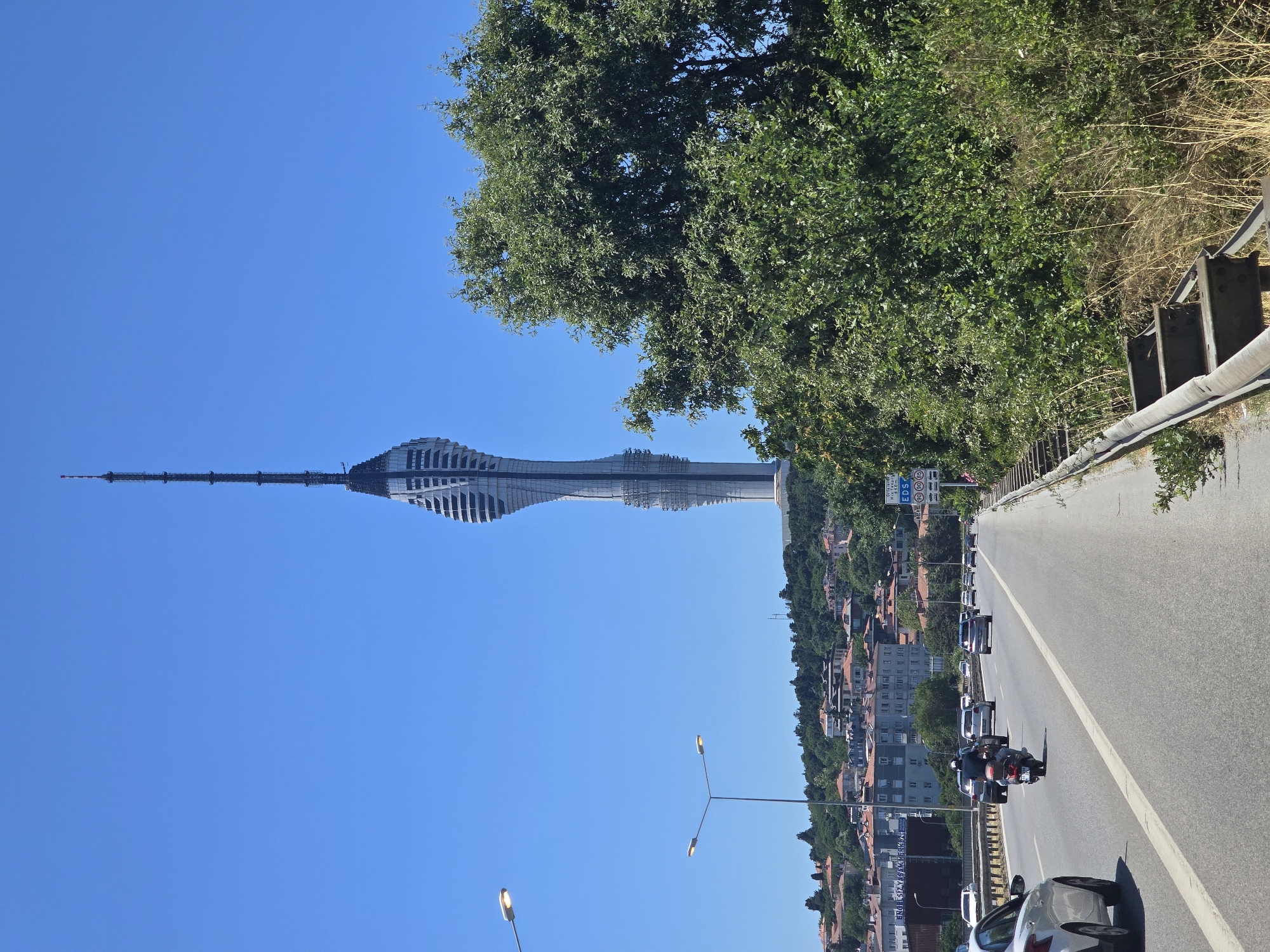 The image captures a view of the Çamlıca Tower, a telecommunications tower located in Üsküdar, Istanbul, Turkey. The tower is a prominent, futuristic-looking structure with a distinctive twisting design and a tall antenna spire, set against a clear blue sky. In the foreground, a multi-lane highway is visible with several cars and a motorcycle in motion. The highway is bordered by a metal guardrail and a dense green tree on the right side of the frame. On the left side of the highway, a residential area with buildings and trees is nestled on a hill. Streetlights are also visible along the highway. The overall scene depicts a sunny day in a city environment.