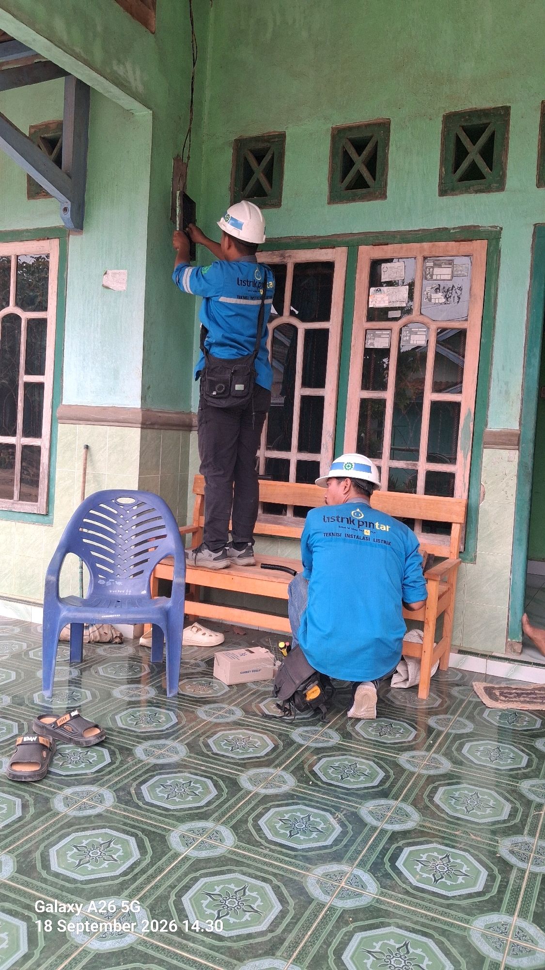 Two individuals, identified by their blue uniforms and hard hats, appear to be performing electrical work on the exterior of a building. The building has light green walls and wooden window frames. One individual is standing on what appears to be a stool, working on an electrical box mounted on the wall. This individual is wearing a blue uniform with "Listrik Pintar" and "Teknisi Instalasi Listrik" written on the back. They are also wearing dark pants and a black shoulder bag. The second individual is sitting on a wooden bench, facing away from the camera and towards the building. This individual is also wearing a similar blue uniform with "Listrik Pintar" visible on the back. They are wearing dark pants and a backpack. In the foreground, there is a blue plastic chair, a pair of sandals, and a small cardboard box. The ground is covered in patterned green tiles. The image is dated "18 September 2026 14:30". The location is identified as Karang Anyar, Indonesia.