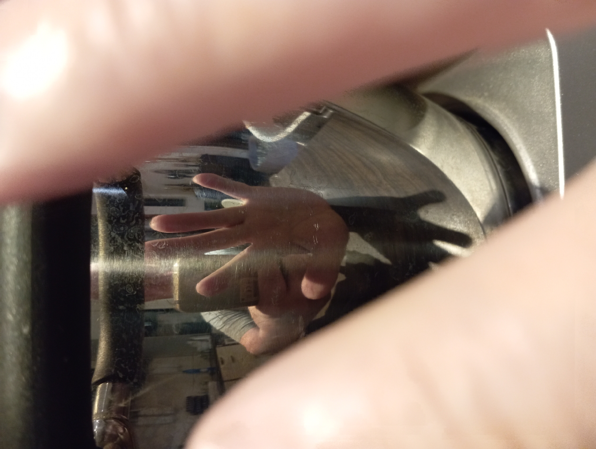 The image is a close-up, abstract shot of a person's hands interacting with a reflective surface, possibly a polished metal object like a coffee machine or a car part. The foreground is dominated by blurred, fleshy tones of what appear to be fingers and palms, obscuring much of the scene.
Reflected in the surface, we can see a distorted view of a hand, with fingers splayed, reaching out. The reflection also captures a glimpse of the person's arm and torso, wearing a grey t-shirt. The background within the reflection shows a domestic or workshop setting with some indistinct objects and furniture, hinting at an indoor environment. The lighting suggests it's daytime, with a bright light source creating highlights on the reflective material and casting shadows of the hand.
The overall impression is one of tactile exploration and curiosity, perhaps a moment of someone inspecting or using the object. The abstract nature of the shot, with its emphasis on texture and reflection, creates a sense of intimacy and focuses the viewer's attention on the interaction between the human form and the material world. There is no visible text in the image. The location context of Baden, Austria, does not provide specific visual cues within the image itself.