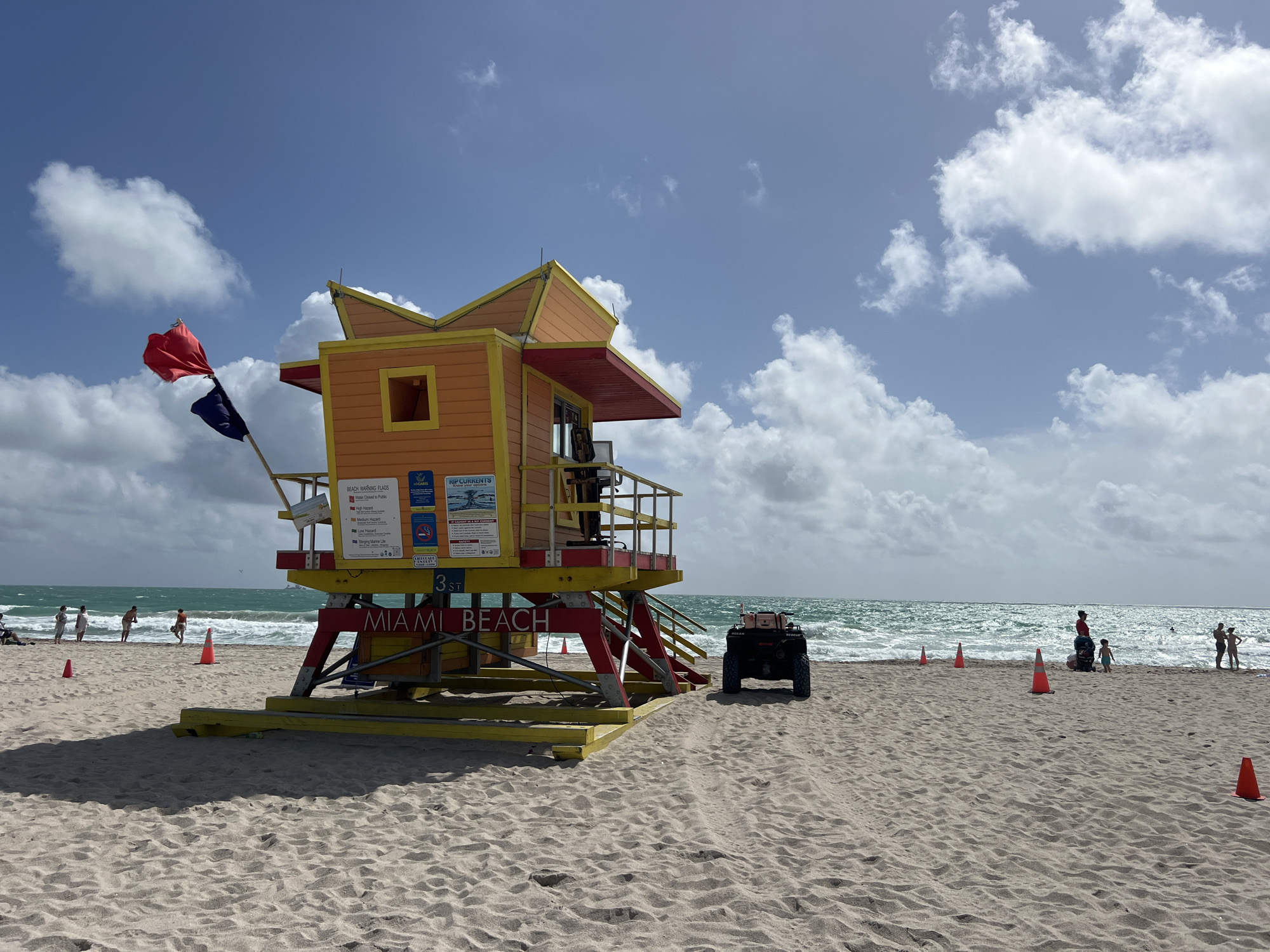 The image depicts a lifeguard station at 3rd Street, Miami Beach, United States of America, on a sunny day with scattered clouds. The station is painted in orange, yellow, and red colors. A red flag and a dark blue flag are flying on the left side of the station. Signage is present, with text including "BEACH WARNING FLAGS" and "RIP CURRENTS". In front of the station, on the sand, sits a black all-terrain vehicle (ATV). Orange traffic cones are sparsely scattered on the beach. In the background, the ocean meets the horizon, and people are visible wading in the water or standing on the shoreline.