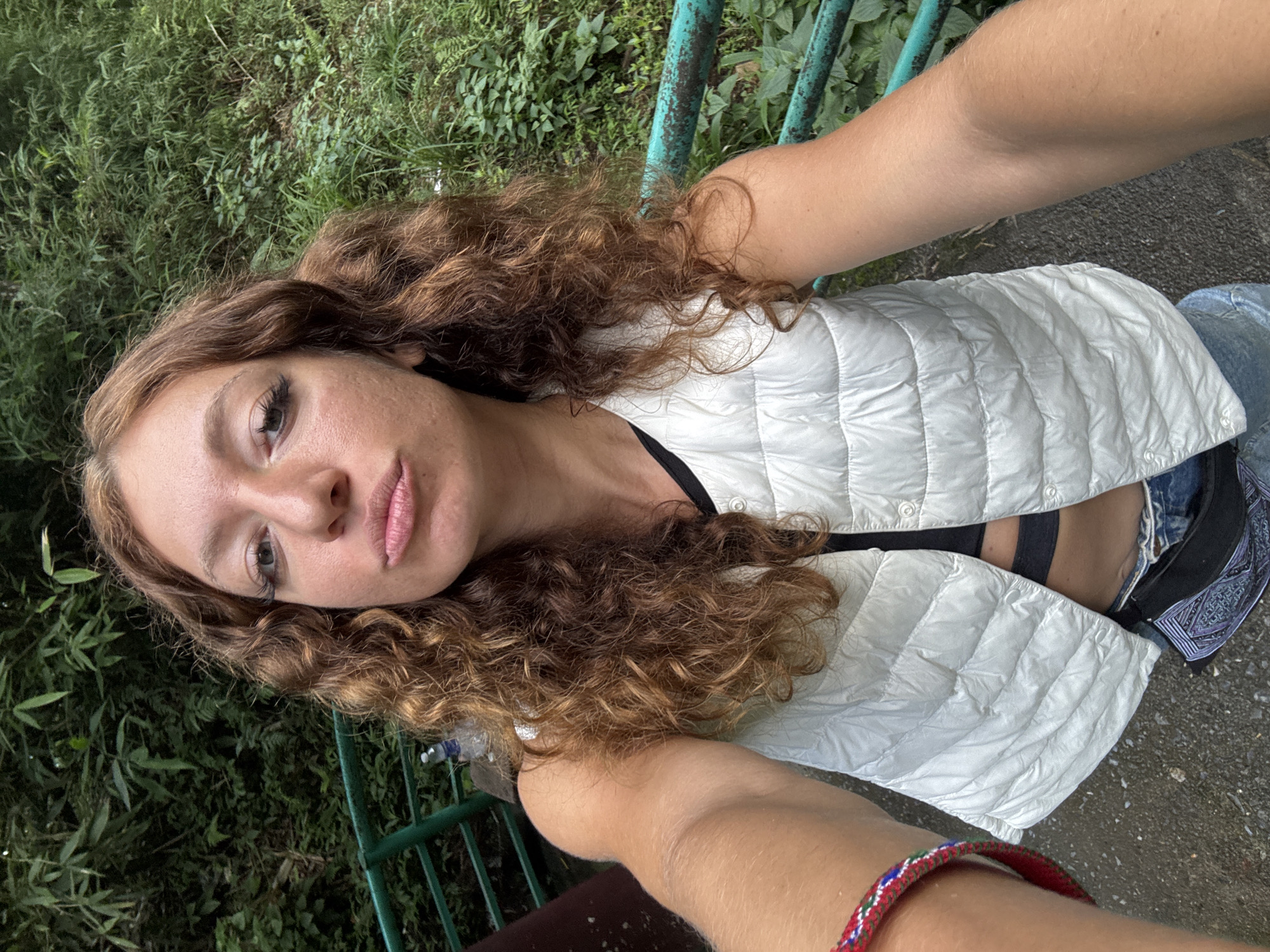 A young woman takes a selfie outdoors. She has curly, reddish-brown hair, and is wearing a white quilted vest over a black top and denim shorts. A section of the vest is open, revealing a portion of her stomach.
The background consists of lush greenery and a turquoise metal railing. There is a section of pavement visible in the lower right. Her left arm, adorned with a red beaded bracelet, stretches toward the camera. The location is Sa Pa Ward, Vietnam.