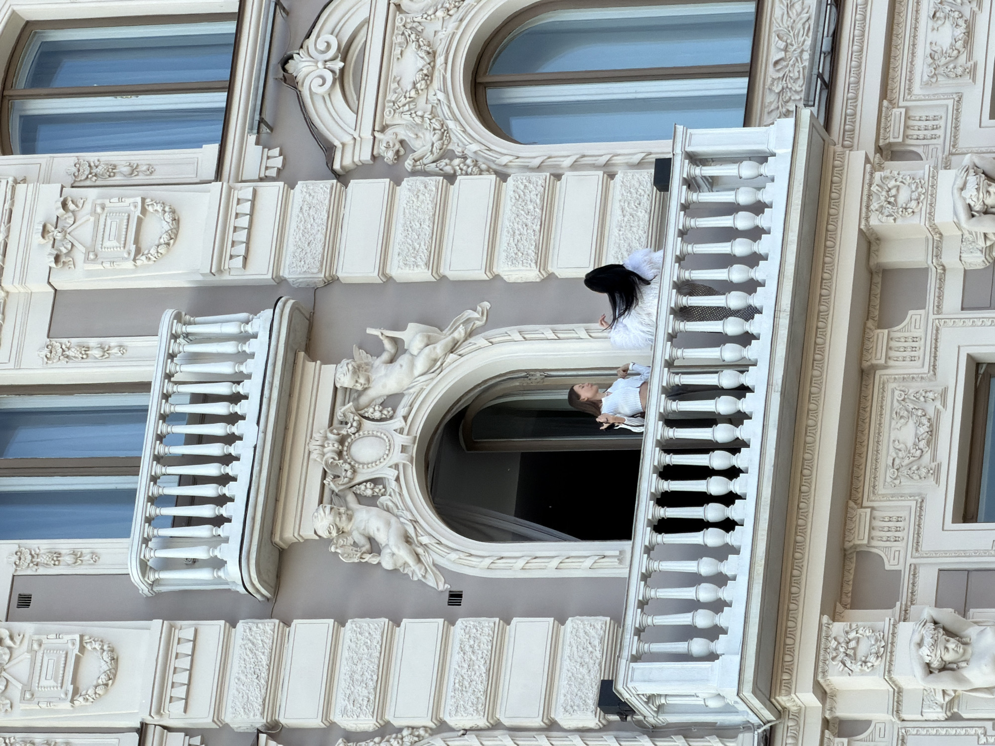 A building facade in Saint Petersburg, Russia, is visible. The facade is adorned with decorative elements, including cherub statues and ornamented window frames. A woman wearing a white outfit and a black feathered headdress stands on a balcony. A second person is looking out from a window or balcony beside her. The architecture is classical in style, with pale colors and intricate details. The photograph appears to be taken from a ground-level perspective, looking upward at the building.