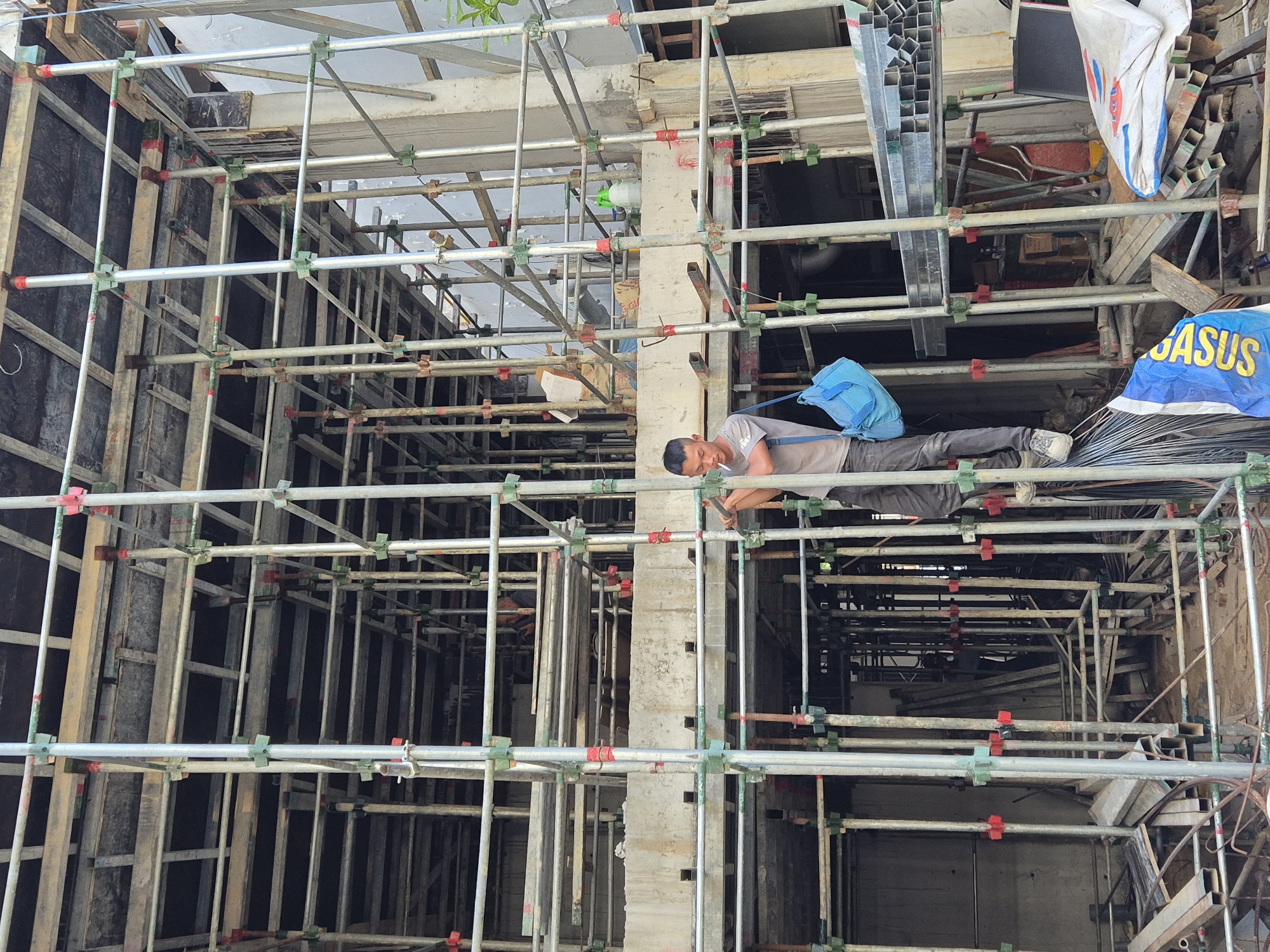 A construction worker, dressed in a gray t-shirt and dark pants, stands on scaffolding at a building site in Đà Nẵng, Vietnam. He is holding a tool and appears to be smoking a cigarette. A blue bag hangs from his shoulder. The scaffolding is made of metal poles and connectors, with wooden planks forming the walkways. The building under construction is visible in the background, with concrete structures and exposed framework. Various construction materials, including metal pipes and bags of cement, are scattered around the site. The scene depicts a typical day on a construction project in an urban environment.