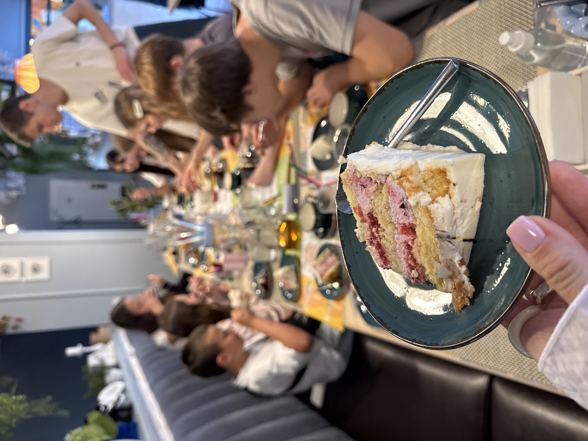 The image captures a group of people gathered around a long table, likely enjoying a meal together. The setting appears to be indoors, possibly in a restaurant or dining establishment. The focus is on a slice of cake, featuring layers of sponge, a pink filling (possibly raspberry), and topped with white frosting. The cake rests on a dark teal plate. A hand with manicured pink nails holds the plate. Numerous other people are seated along the table; some are actively engaged with their food or drinks, and others are in conversation. The overall atmosphere seems convivial and lively. The location in the city of Sochi, Russia, cannot be determined from the provided image and information.