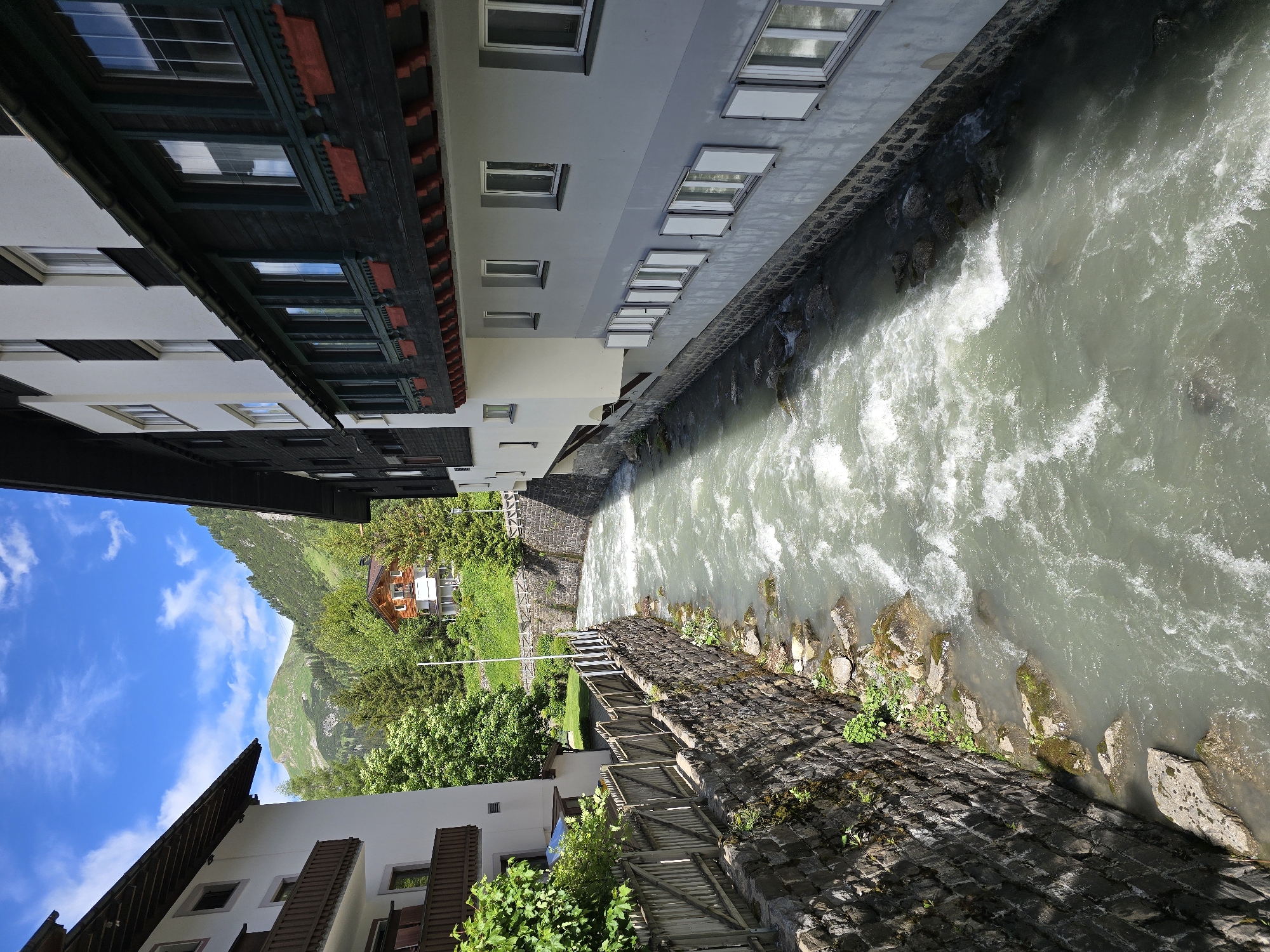 The image depicts a view of the Lech River flowing through the town of Lech, Austria. The river, with its turbulent, greenish water, is bordered by stone retaining walls and wooden fences. On the left side of the river, a white building with wooden balconies is visible, featuring lush green foliage. On the right side, a multi-story building with white and gray facades and numerous windows is prominent. The windows on the lower levels have white shutters, some of which are open. The upper parts of the buildings are dark-colored, with decorative wooden elements and red flower boxes. In the background, a green mountain slope rises, dotted with trees and a small red-roofed house. The sky is blue with scattered white clouds, suggesting a clear day. There are no people visible in the image. The scene captures the natural beauty of the alpine environment combined with the architecture of the town.