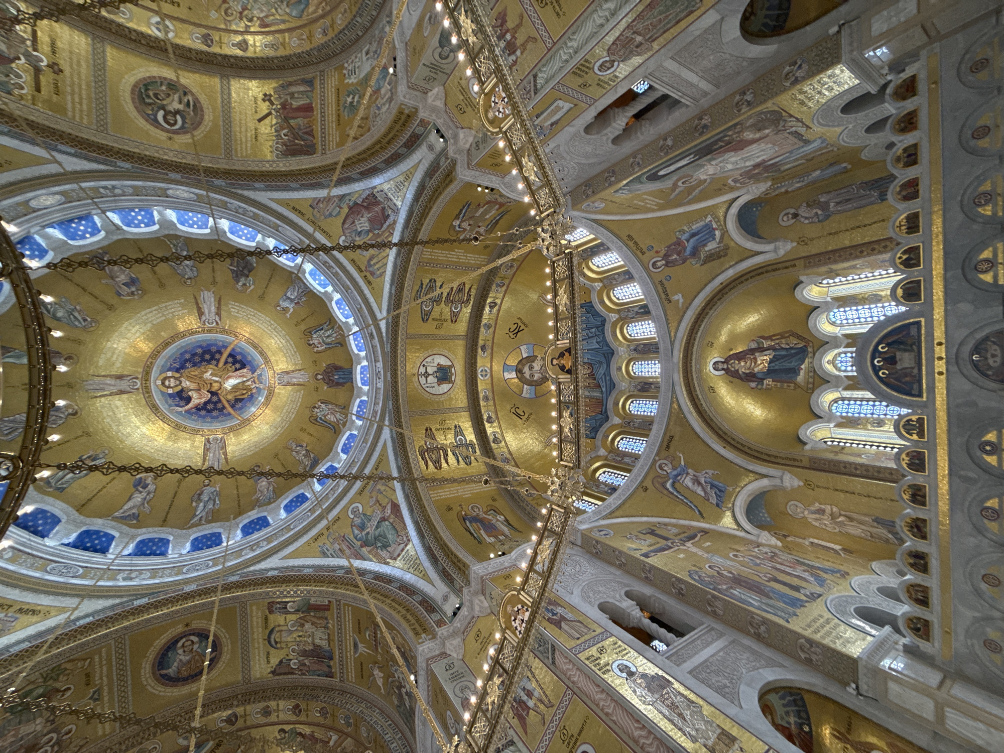 The image captures the majestic interior of a large Eastern Orthodox church, showcasing an elaborate ceiling adorned with intricate golden mosaics. The perspective is looking directly upwards, revealing multiple domes, arches, and vaulted sections.
The primary subjects are numerous religious figures and biblical scenes rendered in mosaic. The central dome features a depiction of Christ, possibly as Pantocrator, surrounded by saints or apostles, while other domes and arches display a multitude of saints, angels (such as "ГАВРИЛЬ" - Gabriel), and narrative scenes. One identifiable scene is "Біговіщеніе ПРЕСТЫНА БЦЫ" (Annunciation of the Most Holy Theotokos). Figures are depicted in traditional Byzantine iconography, characterized by stylized forms and serene expressions.
The dominant material is glittering golden mosaic tiles, creating a radiant and opulent effect throughout the ceiling. Sections of the domes and arches also incorporate blue and white mosaics, particularly around windows, adding a vibrant contrast. Ornate chandeliers, adorned with numerous small lights, hang from the ceiling, casting a warm glow that enhances the shimmer of the golden tiles. Natural light also filters through arched windows set into the structure.
Visible text in Cyrillic script identifies many of the figures and scenes, including "СТ МАРКО" (Saint Mark), "ЖВАНЂЕЛЕКТ MATEJ" (Evangelist Matthew), "свети Апостол" (Holy Apostle), and the Christogram "IC XC". Other partial words like "CBCTH" (Holy/Saint) are also discernible.
The scene depicts the highly decorative and revered interior of a place of worship, likely a cathedral, in Belgrade, Serbia, emphasizing its rich spiritual and artistic heritage. The overall impression is one of grandeur, sacredness, and meticulous artistry, bathed in a warm, inviting light.