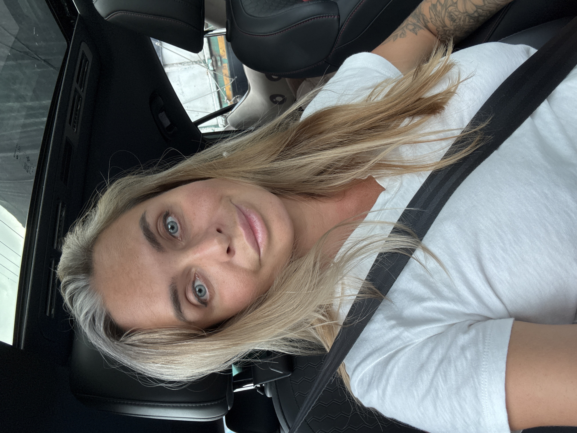 A blonde woman with blue eyes is taking a selfie in the driver's seat of a car. She is wearing a white t-shirt and a black seatbelt. Her hair is long and blonde, with some gray streaks. The car's sunroof is open, and the sky is visible. The interior of the car is dark, with black leather seats. There is a tattoo on her right arm. The image does not provide any information about the location in the city of Pran Buri, Thailand, or any specific context.