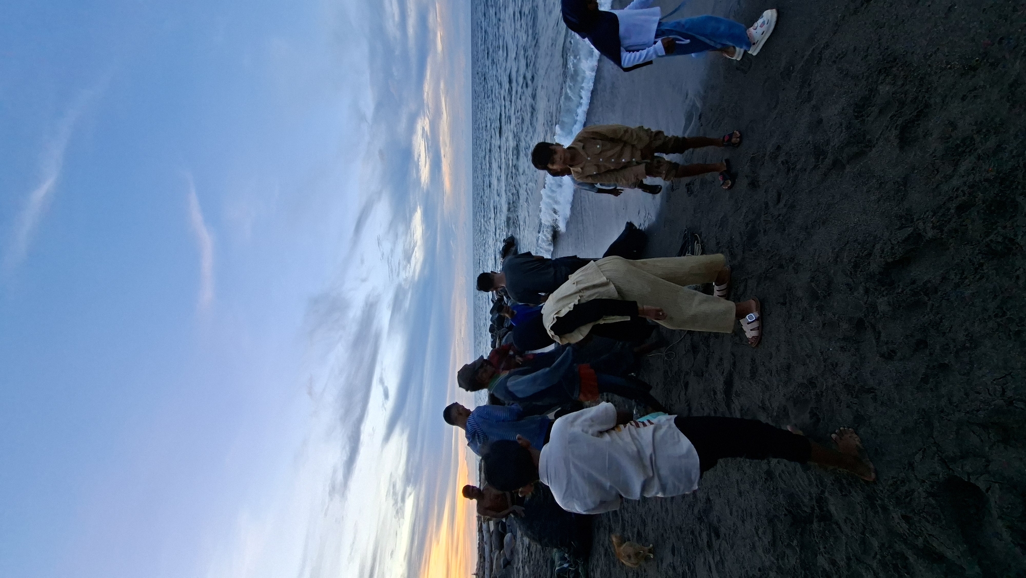 This image depicts a group of people on a dark sand beach near the ocean in Lhokseumawe, Indonesia, during what appears to be twilight. The sky is a blend of pale blue and muted orange hues, with scattered clouds. The ocean waves are gently reaching the shore.
In the foreground, a young boy in a white shirt and black pants stands with his back to the camera. To his right, a person in a tan outfit, possibly a woman, is bent over. Further right, a young boy in a tan shirt and shorts is looking towards the viewer, and a young girl in a blue headscarf and white shirt is visible. Several other individuals, including men and children, are scattered throughout the scene, some near a pile of dark objects on the sand that could be fishing nets or debris. A small, light-brown dog is present on the sand to the left. The overall atmosphere is one of casual gathering or activity by the sea.