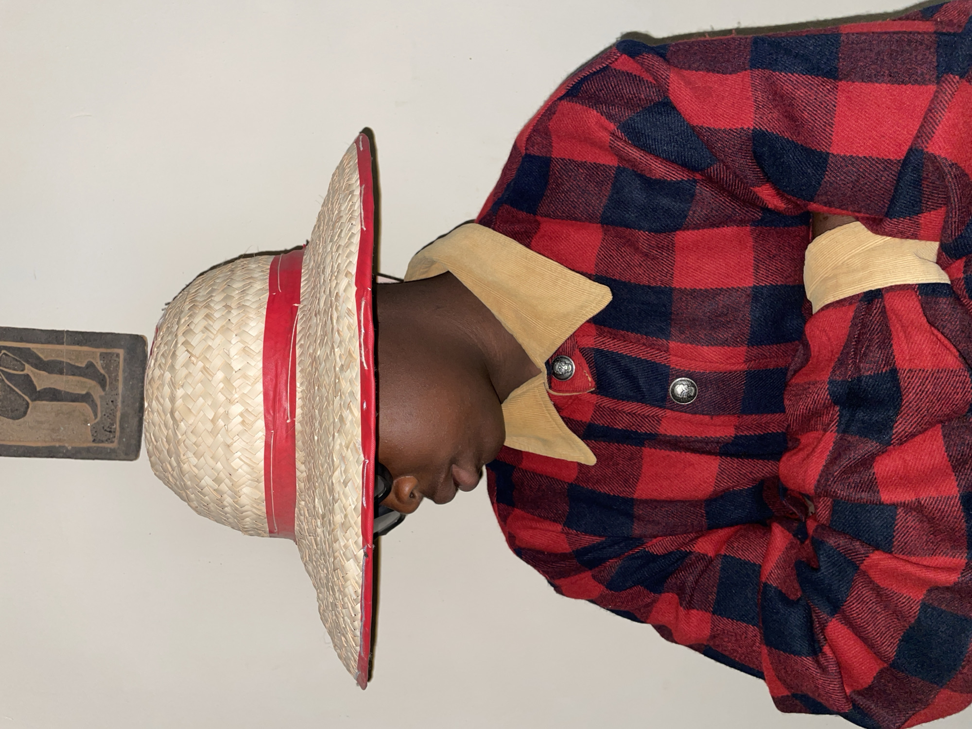 A person is shown wearing a red and black plaid flannel shirt with a tan corduroy collar. They are also wearing a straw hat with a red band around the brim. The background is a white wall. There is a decorative tile on the wall to the left of the hat. The location is not mentioned.