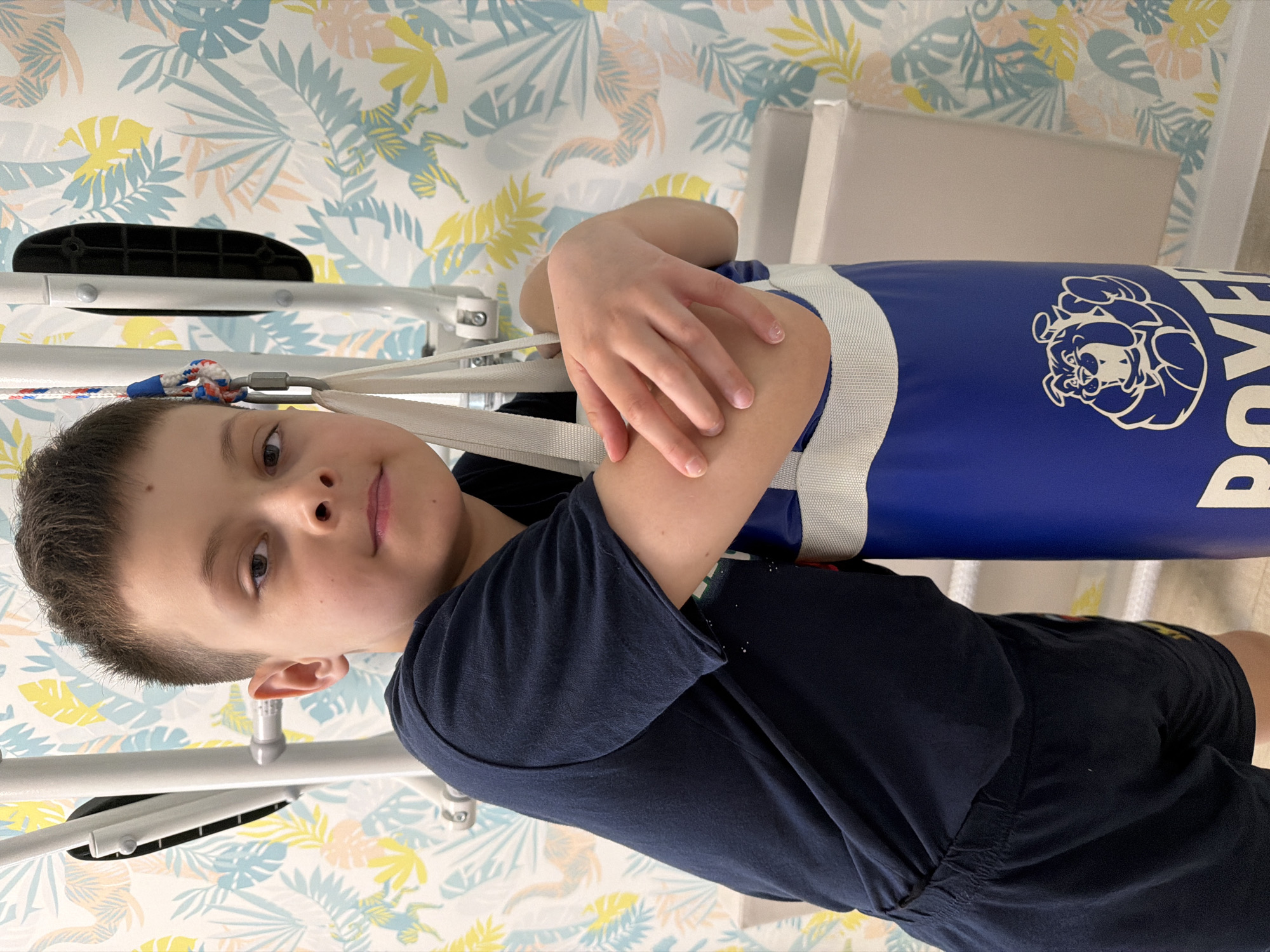 A boy stands indoors next to a blue punching bag, which hangs from a white metal frame. The boy wears a dark blue t-shirt and dark shorts. He has short brown hair and appears to be looking at the camera. His right arm is bent and positioned across the top of the punching bag. The background is a wall with a yellow, blue, and peach leaf patterned wallpaper. The location is Votkinsk, Russia.
