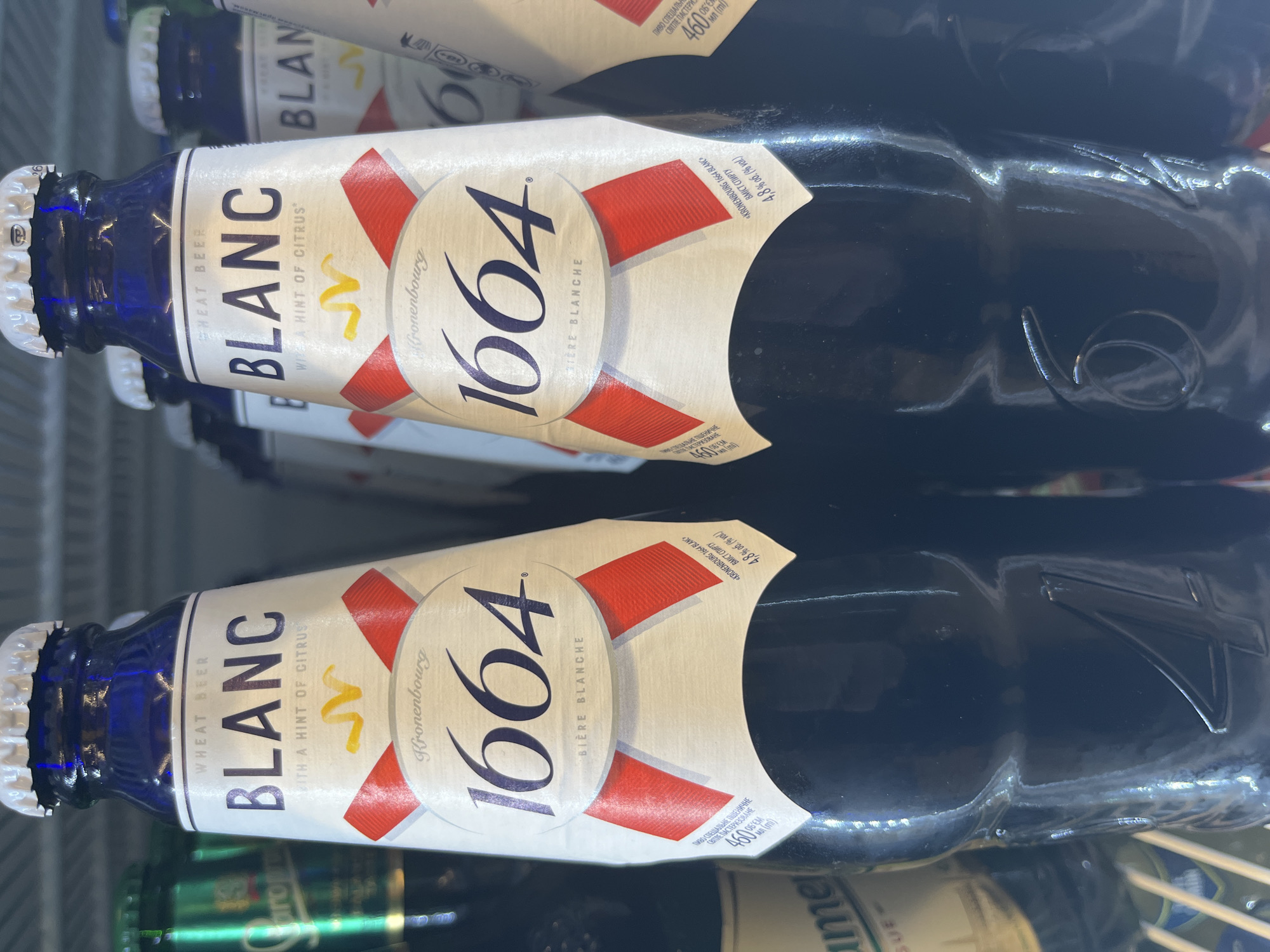 This image shows two bottles of Kronenbourg 1664 Blanc wheat beer in a cooler or refrigerator. The bottles are dark blue glass with white labels featuring red and white diagonal stripes and a stylized "K" logo. The text on the labels indicates "WHEAT BEER BLANC WITH A HINT OF CITRUS" and "BIÈRE BLANCHE." Some Cyrillic text is also visible on the labels, including "ПИВО СПЕЦІАЛЬНЕ ПШЕНИЧНЕ" (Special wheat beer) and "4,8% об. (% vol.)" (4.8% vol.). The image is taken from a slightly angled, close-up perspective, with the bottles oriented vertically. The lighting suggests an indoor setting, likely a retail environment where the beer is being sold. The reflections on the bottles indicate a smooth, glossy surface. The overall impression is a product shot focused on the branding and details of the beer bottles.