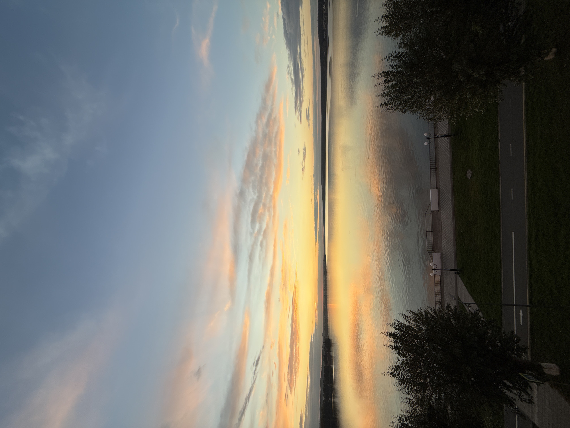 This is a high-angle, wide shot of a lake at sunset in Votkinsk, Russia. The sky is a gradient of light blue and soft orange and pink hues from the setting sun, with wispy clouds scattered throughout. The calm lake perfectly reflects the colors of the sky, creating a mirror-like surface. In the foreground, a paved promenade runs along the edge of the lake, bordered by a low railing. Adjacent to the promenade is a strip of green grass, and beyond that, a road with a white lane marking and an arrow indicating direction. Two trees, one on the left and one on the right, partially frame the scene. A lamppost with two lights stands on the promenade. The distant shore of the lake is visible as a dark, low-lying silhouette. No people are visible in the image.