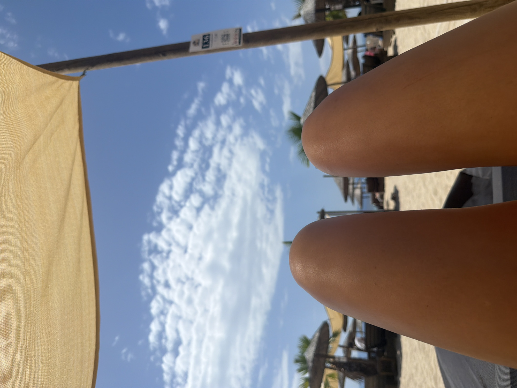 The image shows a first-person perspective of a person's legs on a beach in Ofrynio Beach, Greece. The legs are tanned and are resting on a dark surface, likely a beach chair or lounger. The background is a bright blue sky with scattered white clouds. Above the legs, a light tan beach umbrella is visible, creating shade. In the distance, there are more beach umbrellas, palm trees, and beach chairs, indicating a populated beach setting. A wooden pole with a small sign attached to it is also visible on the right side of the image. The overall impression is one of relaxation and enjoying a sunny day at the beach.