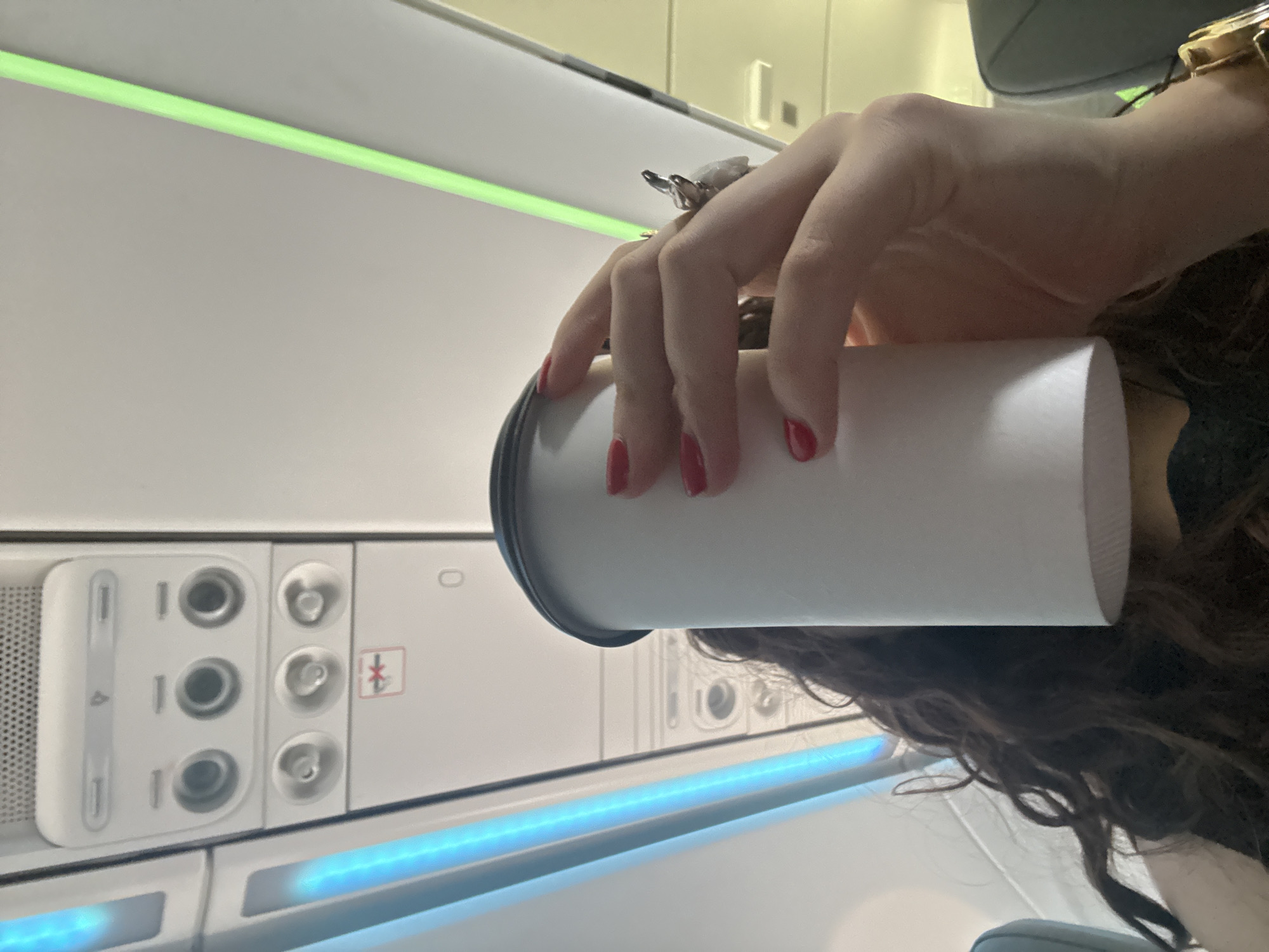 The image appears to be taken inside an airplane. A person is holding a white cup with a dark gray lid. The person has red nail polish, a silver-colored ring, and a gold-colored watch. The person has brown curly hair. The interior of the airplane cabin is visible, including an overhead panel with controls for lights, air, and Bluetooth. There is a blue light strip above the panel. The location in Apodaca, Mexico is not evident from the image.
