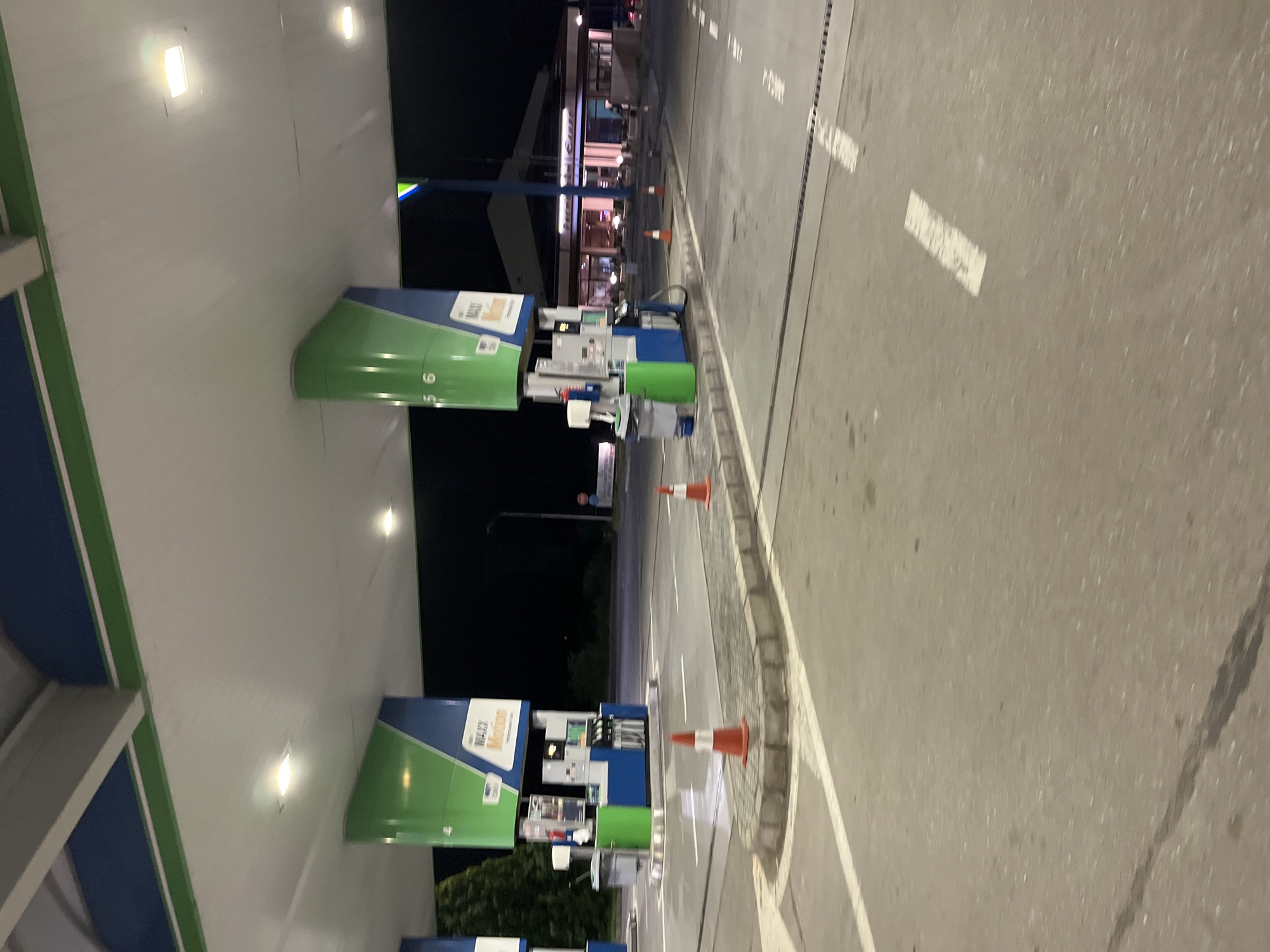 The scene depicts a gas station at night. Visible are multiple gas pumps under a well-lit canopy with blue and green accents. The pumps are marked with numbers, indicating their station number. Orange traffic cones are strategically placed around the pumps, suggesting some form of maintenance, construction, or hazard management.
Beyond the gas station, there is a building with illuminated windows, possibly a restaurant or store. The street adjacent to the gas station has white dashed lines indicating traffic lanes and solid white lines defining the edge of the road.
The environment is likely urban or suburban, indicated by the buildings and street infrastructure. The location may be Popovitsa, Bulgaria. There are no people interacting in the scene. The time is night, with ambient lighting coming from the gas station and surrounding buildings.