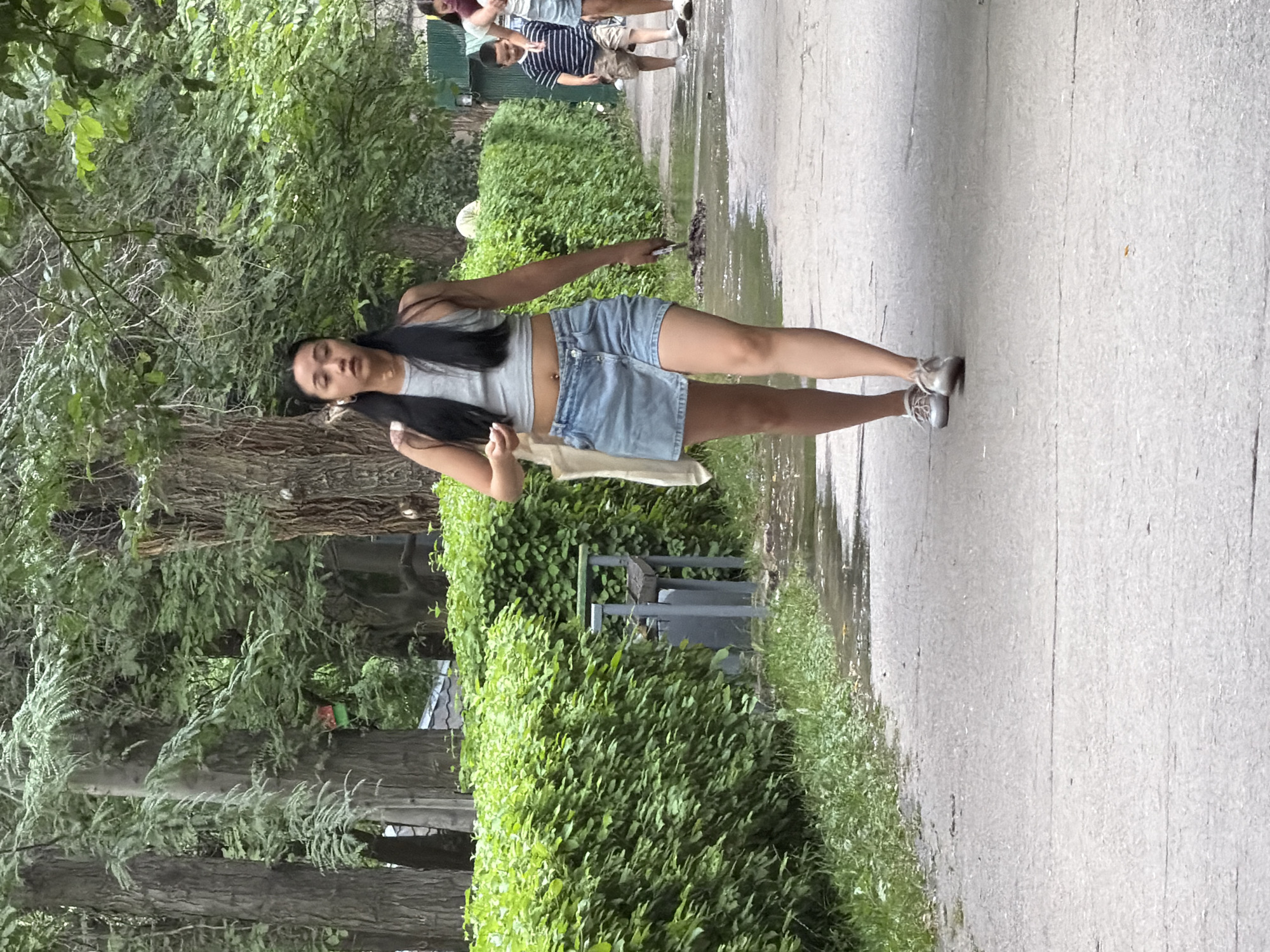 A young woman with long dark hair walks on a paved path in a park. She is wearing a gray crop top and a denim skirt. A white tote bag hangs from her left shoulder. She is looking forward and appears to be holding a phone in her right hand. In the background, a lush green hedge runs along the left side of the path, with trees visible behind it. To the right, a boy in a striped shirt and shorts walks away from the camera, followed by another person. The path is wet in places, suggesting recent rain or watering. A park bench is visible next to the hedge. The overall scene depicts a casual day in a park.