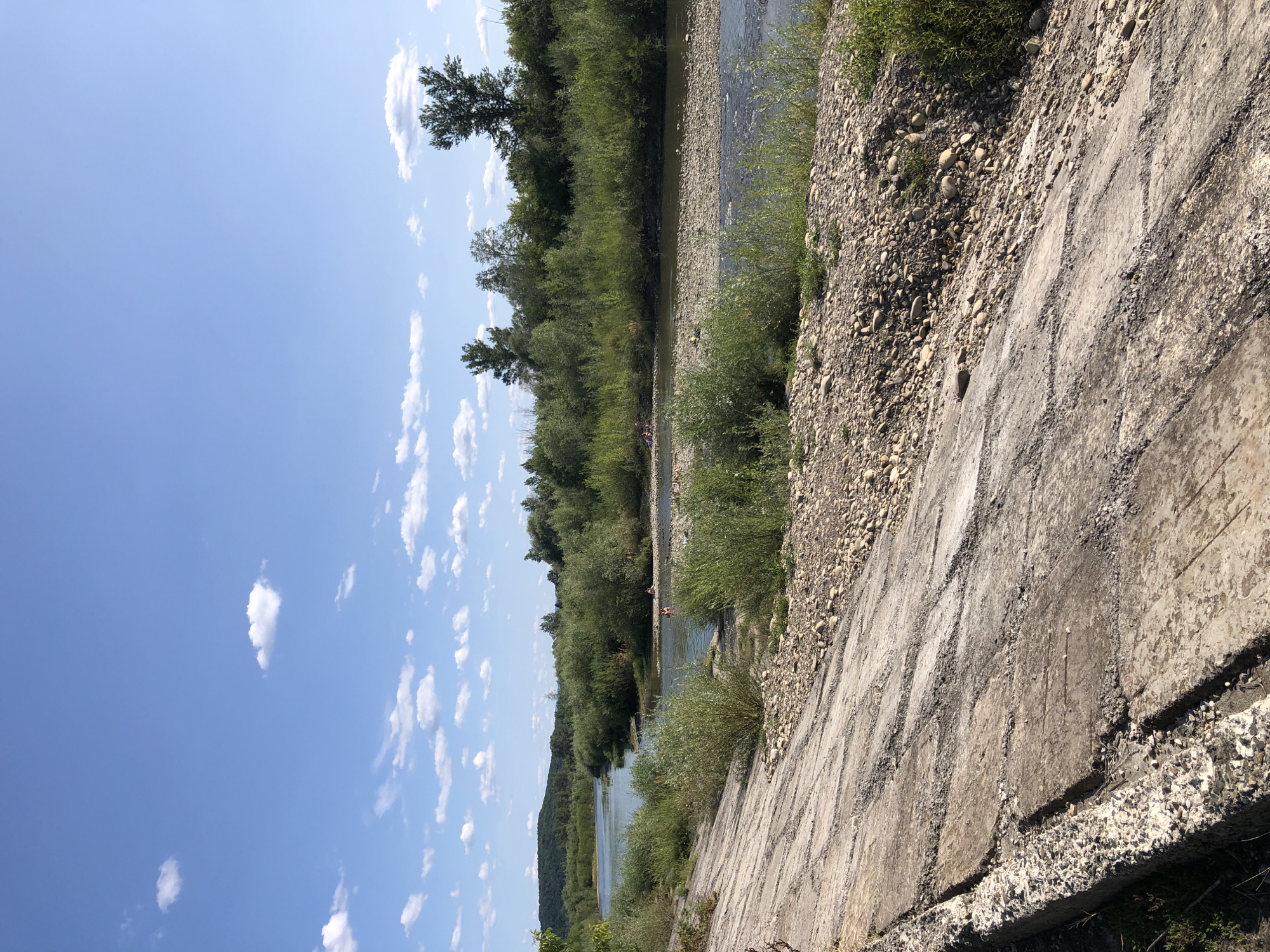 This is a full-shot, landscape-oriented photograph taken outdoors on a sunny day in Nyzhnii Verbizh, Ukraine. The image features a wide, shallow river with a gravelly bed, bordered by lush green trees and bushes on both sides. In the distance, rolling hills covered in trees are visible under a clear blue sky dotted with white, fluffy clouds. In the foreground, a concrete embankment with visible cracks and exposed rebar slopes down towards the river. Several people can be seen in the river, appearing to be swimming or wading.