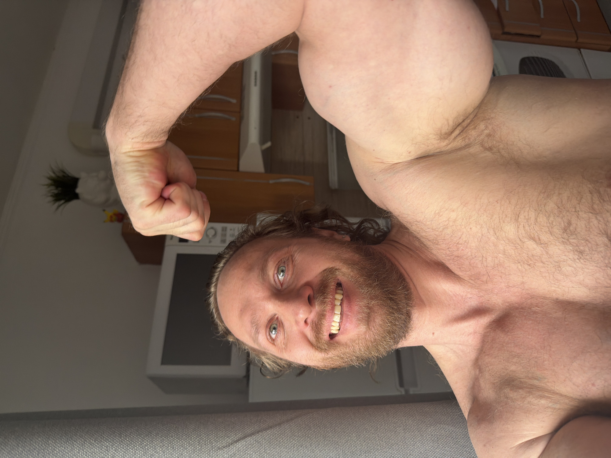 A man with a beard and long hair is posing, flexing his bicep. He is smiling and facing the camera. The background appears to be a kitchen with cabinets, a microwave, and a plant. The location is not identifiable as Saint Petersburg, Russia.