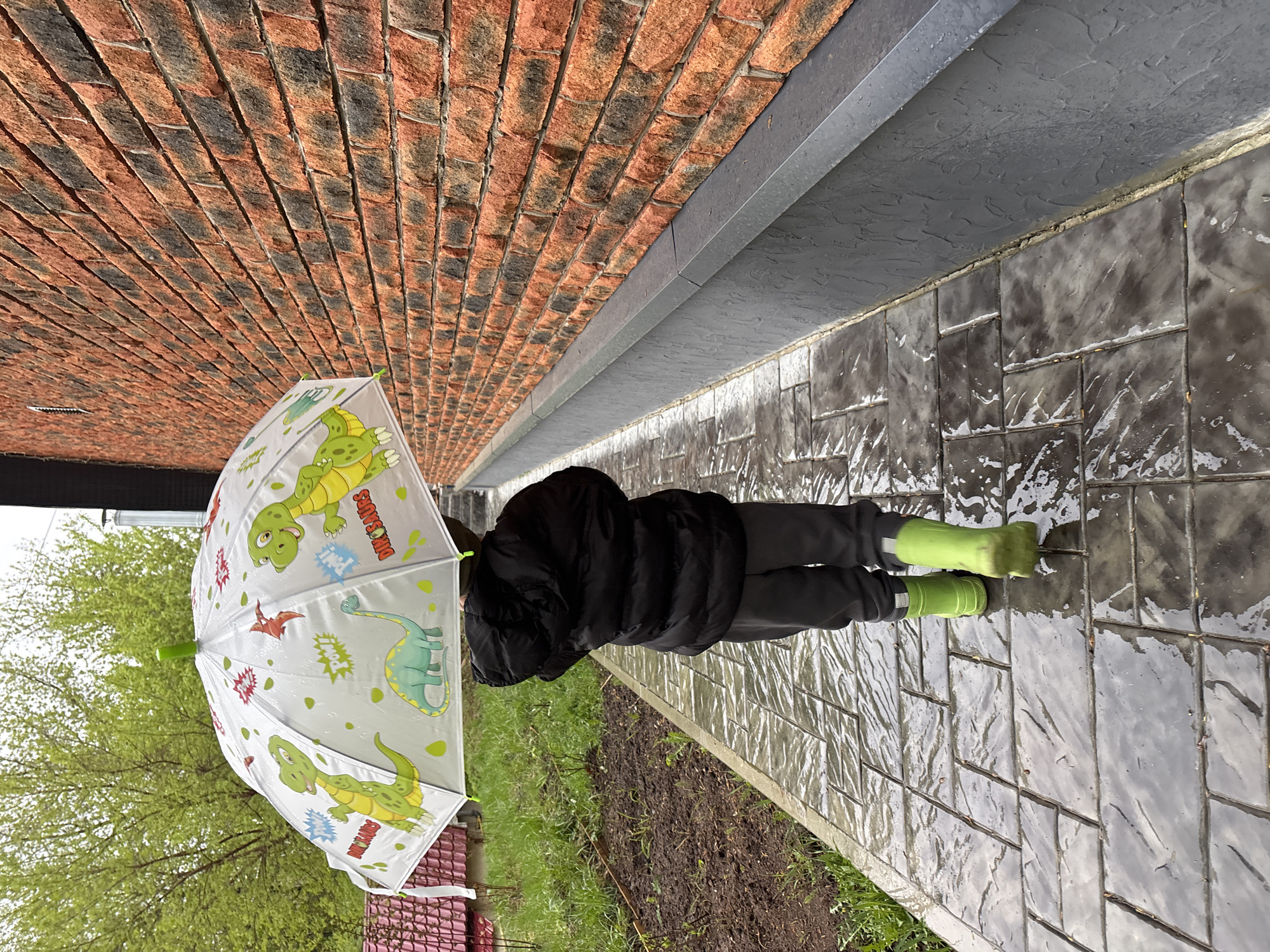 A person in dark clothing and green rain boots stands on a wet, stone-tiled walkway holding a white umbrella decorated with cartoon dinosaurs. A red brick wall is visible behind the person, and bare trees are in the background. Grass and soil are visible alongside the walkway. The location is Chaikovsky, Russia.