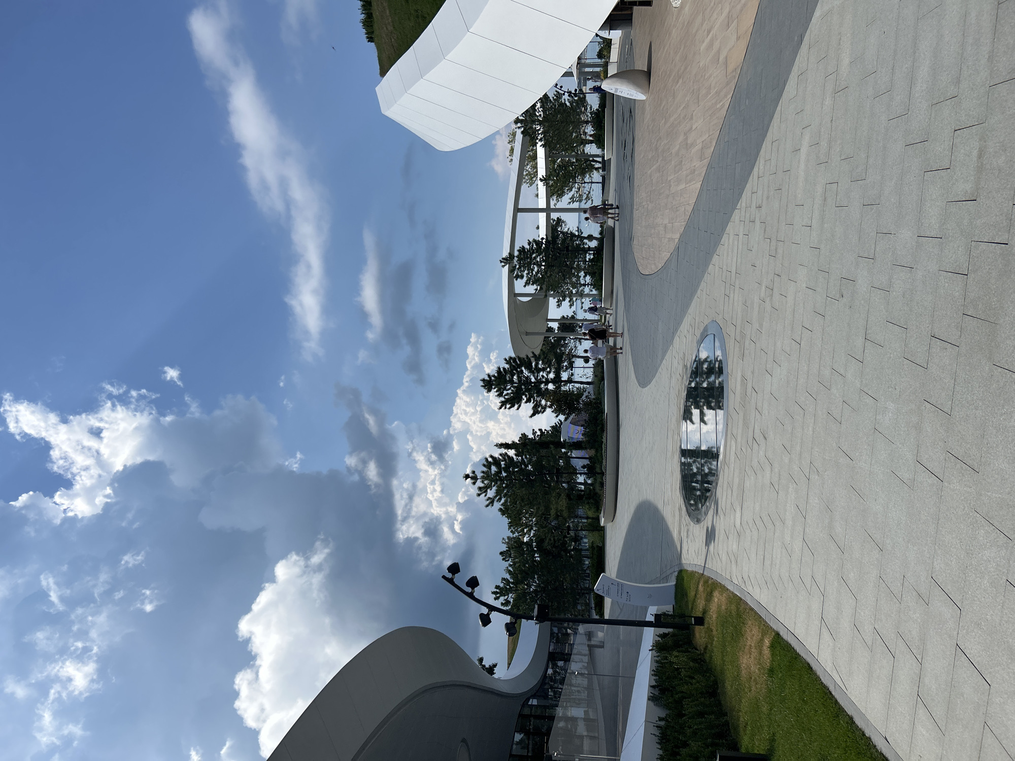 A wide, paved plaza with a modern, curvilinear architectural style is depicted on a sunny day under a partly cloudy sky. The plaza features a light-colored, tiled pathway that curves to the right, leading towards a body of water visible in the background. To the left, a building with a smooth, white, curved facade and large windows is prominent. In the center of the plaza, a circular, reflective pool is embedded in the pavement, mirroring the sky and surrounding trees.
Further into the plaza, several individuals are walking and standing, engaged in casual activities. One of the crops shows a white, abstract-looking structure to the right, resembling a tilted wave or sail. Trees, primarily evergreens, are scattered throughout the scene, with some planted in planters and others forming a small grove. A tall lamppost with multiple light fixtures stands near the building on the left.
The overall impression is a contemporary public space, likely a park or promenade, designed with clean lines and a focus on open areas and natural elements. The location appears to be the embankment in Gelendzhik, Russia, known for its modern waterfront development and public spaces.