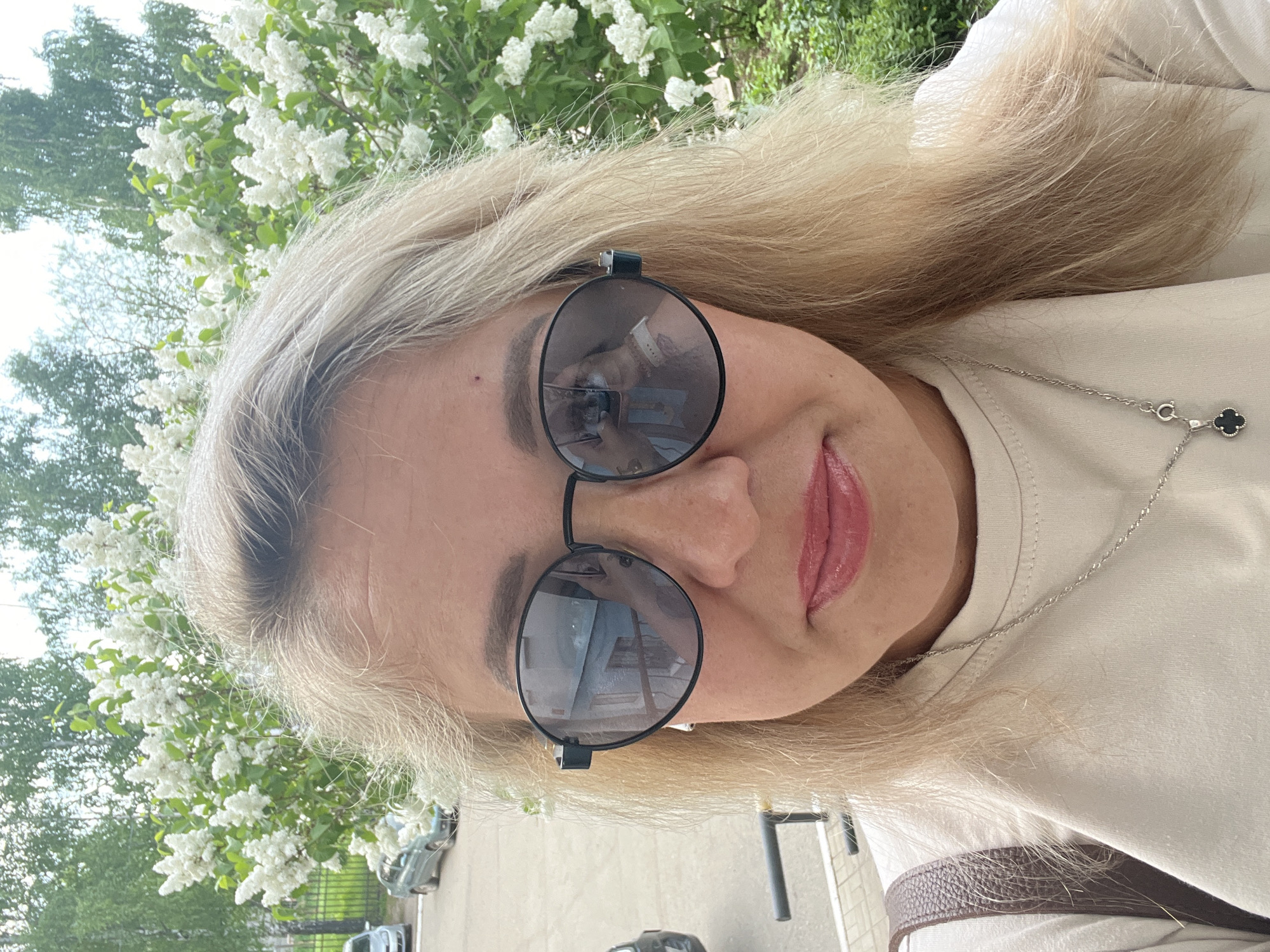 A selfie, showing a blonde-haired person wearing round, dark sunglasses and a light-colored shirt. The person is standing outdoors, with white flowering bushes visible in the background, along with glimpses of parked cars and surrounding green trees. The subject's necklace with a four-leaf clover pendant can be seen, and the reflection in the sunglasses shows a building, which could indicate the location in the city of Chaikovsky, Russia.