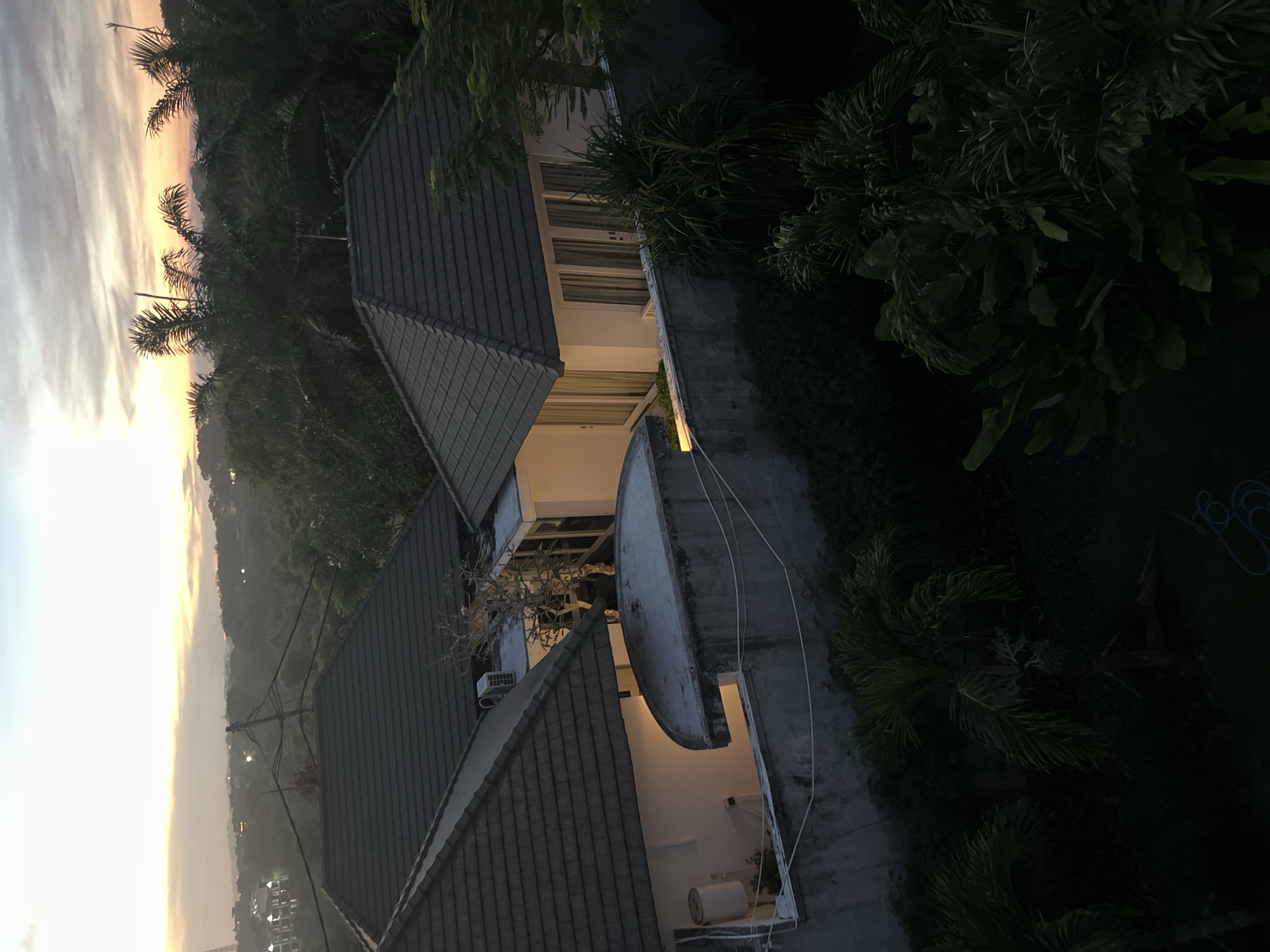 Here is a detailed description of the image:
**Scene:** An elevated aerial view, possibly from a higher building or drone, captures a residential area at either sunset or sunrise.
**Environment:** The sky exhibits a gradient of colors, transitioning from bright yellows/oranges near the horizon to lighter blues and grays further up. The lower portions of the scene show a dense, hilly landscape covered with trees. Closer to the buildings, there is a mix of palm trees and manicured shrubbery.
**Buildings:** Several houses with prominent dark, tiled roofs are visible. The architecture appears modern and features large windows. Some houses are built adjacent to each other, creating a sense of community. One of the houses appears to have a curved or circular structure attached, possibly a pool or a modern architectural feature. The white-painted walls are partially illuminated.
**Objects:** Utility wires traverse the lower part of the image. An air conditioning unit is visible on the side of one of the houses.
**Time:** The light suggests either dawn or dusk.
**Location:** While the specific city and region cannot be determined from the image, the architectural style and vegetation suggest a tropical or subtropical location.