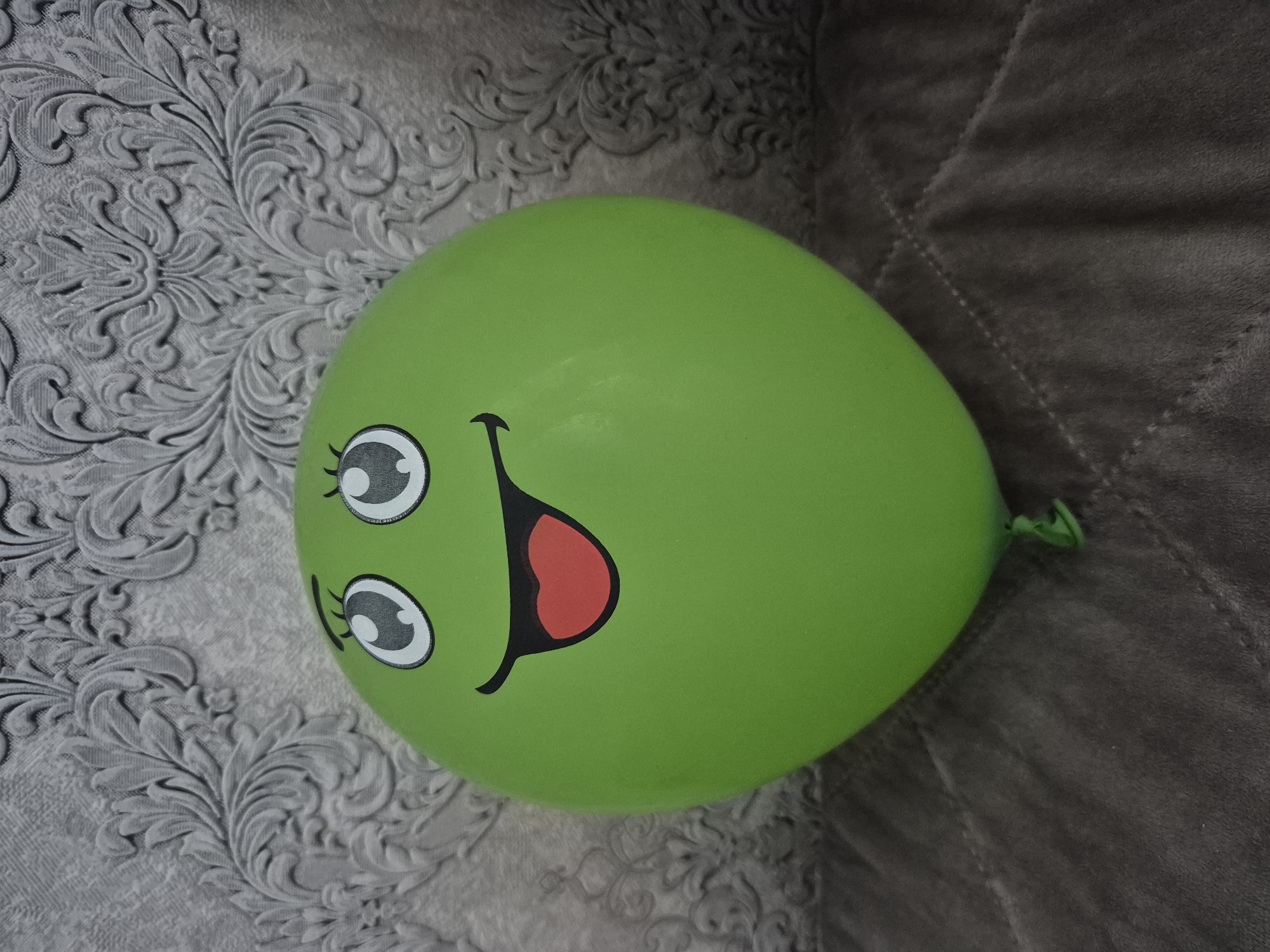 A green balloon with a cartoon face is displayed against a textured gray background. The balloon has large, expressive eyes with black outlines and pupils, and a wide smile with a red tongue sticking out. The background appears to be a wall with a damask pattern, possibly wallpaper. The balloon is resting on a textured surface, likely a blanket or upholstered furniture, which is dark gray and quilted. The image is a close-up shot, focusing on the balloon. There are no people present in the image. The location is not specified in the image, and therefore cannot be determined to be in Astana, Kazakhstan.